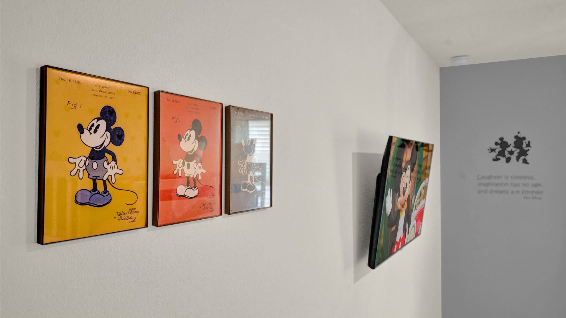 A hallway or common area wall decorated with multiple framed artworks featuring vintage Mickey Mouse patent drawings and a modern Mickey Mouse image. A motivational quote about laughter and dreams is also displayed on the wall.