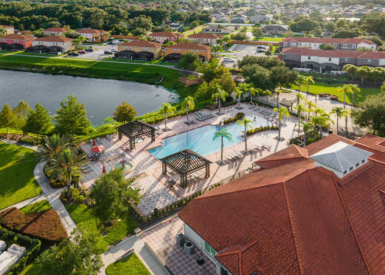 An aerial view showcasing a large communal swimming pool complex with multiple pergolas, surrounded by lush landscaping and palm trees, adjacent to a lake and residential homes.