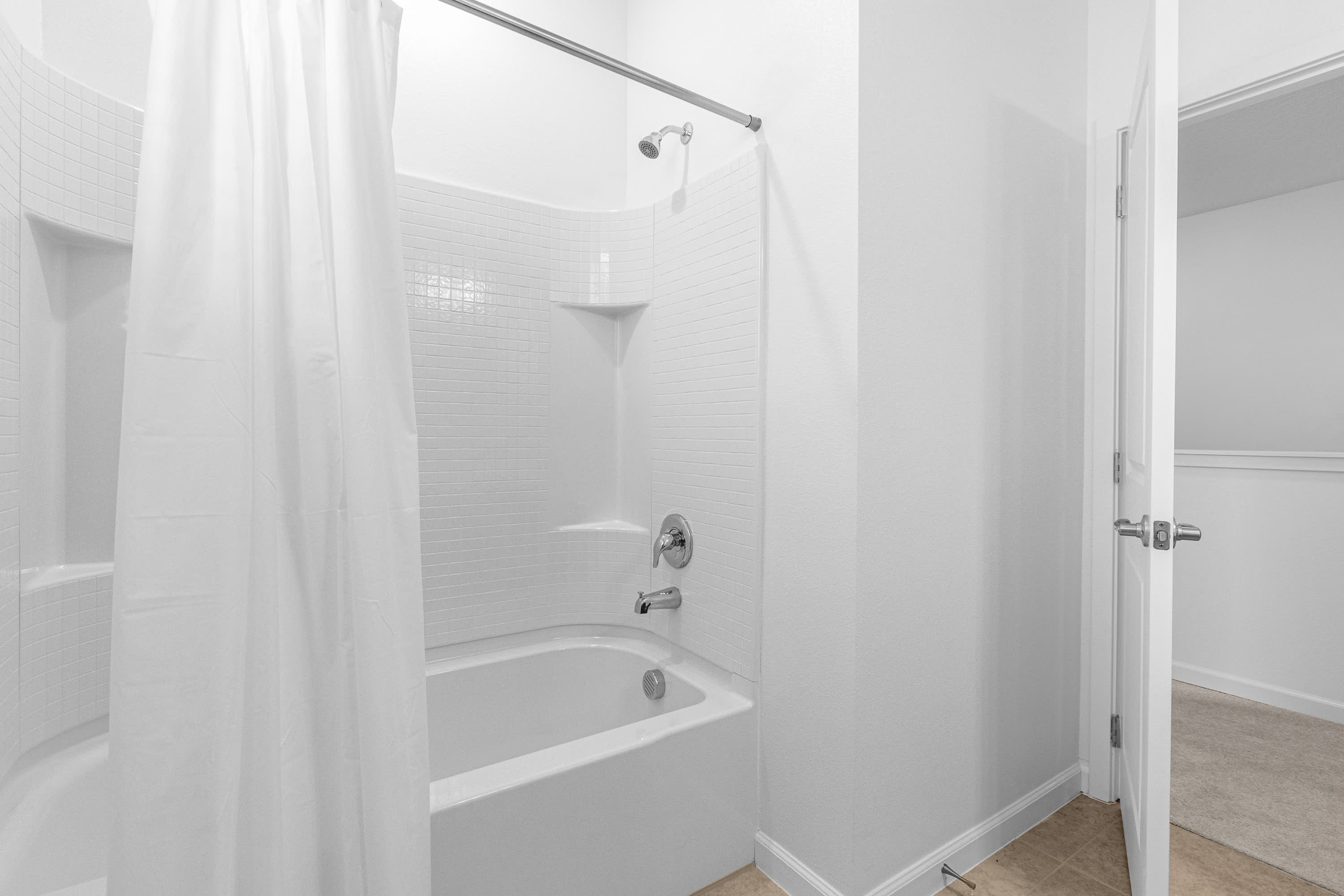 A clean, minimalist bathroom featuring a white bathtub with a shower head and white subway tile surround, enclosed by a simple white shower curtain.
