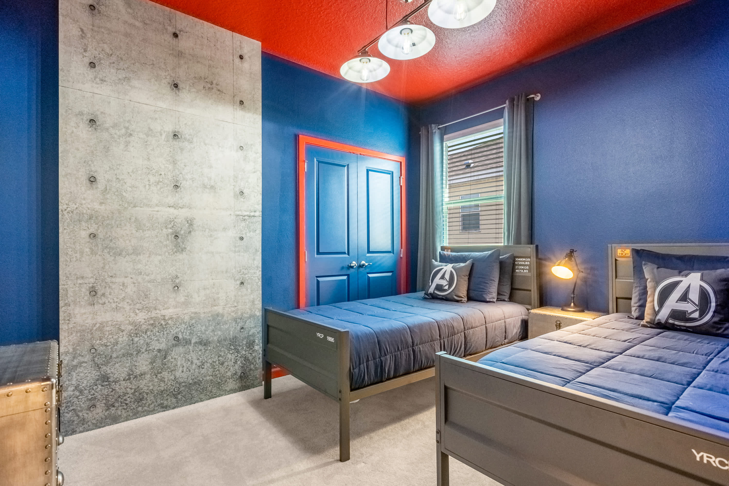 A vibrant themed bedroom featuring two twin beds with dark blue bedding and Avenger-themed pillows, set against bold blue walls, a striking red ceiling, and a concrete-textured accent wall.