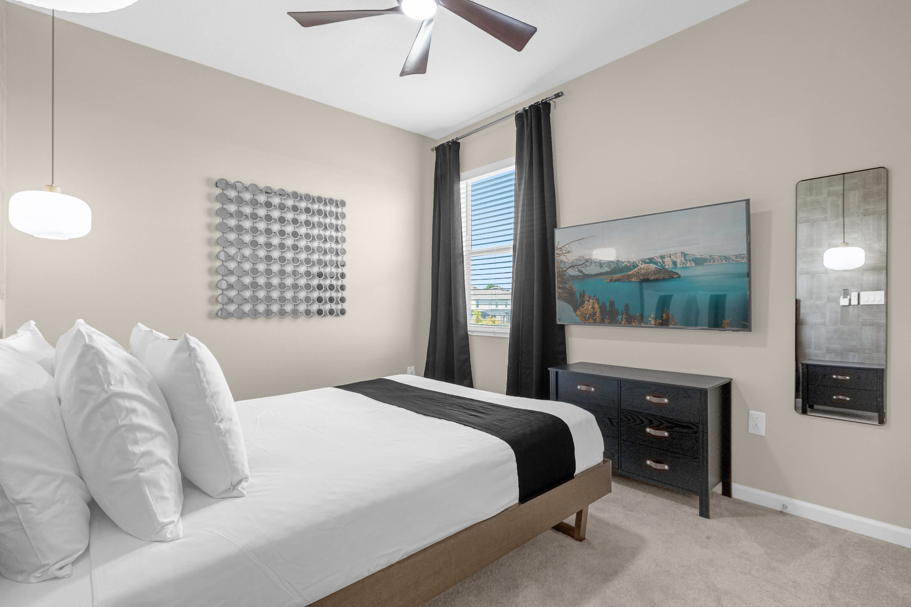 Cozy bedroom with a queen bed, light beige walls, and a wall-mounted TV above a dark dresser. A ceiling fan and black curtains frame the window, with a tall mirror on the adjacent wall.