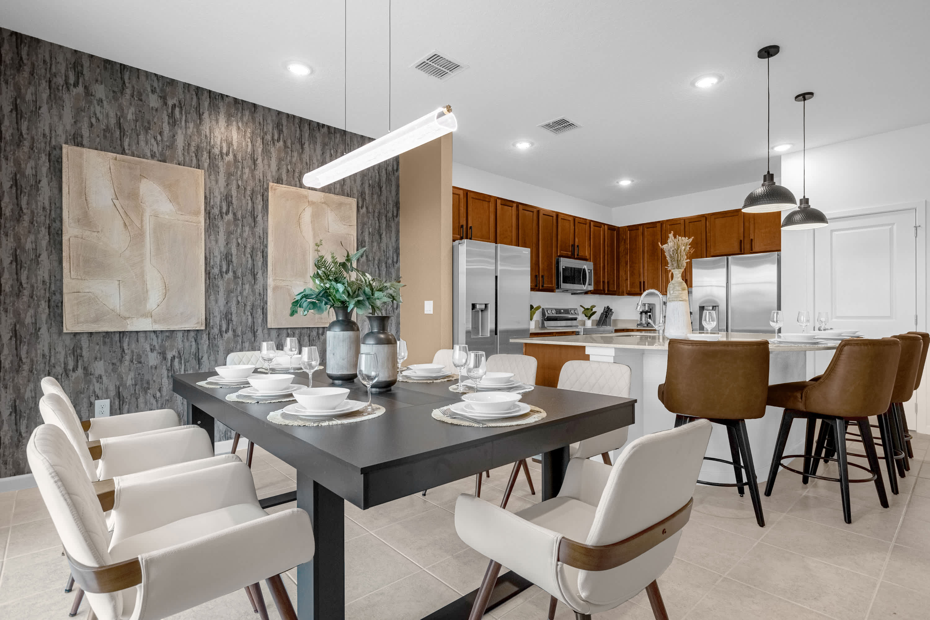 Modern dining area with a dark rectangular table and white upholstered chairs, set against a textured dark grey wallpapered accent wall with abstract art. The open-concept space flows into a kitchen with wood cabinets and stainless steel appliances.