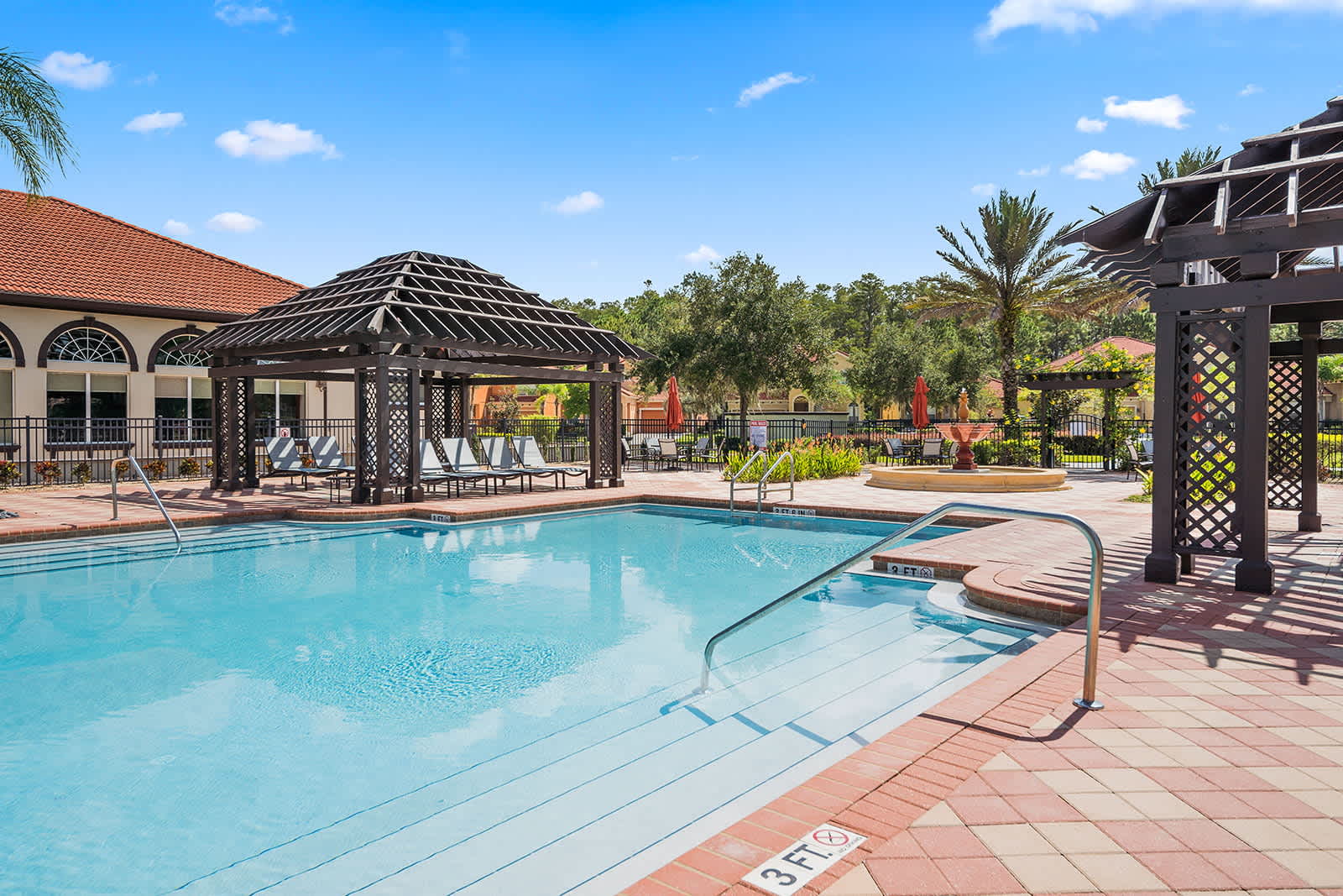 A close-up view of a large, inviting communal swimming pool with clear blue water, featuring steps leading into the shallow end and shaded pergolas with lounge seating nearby.