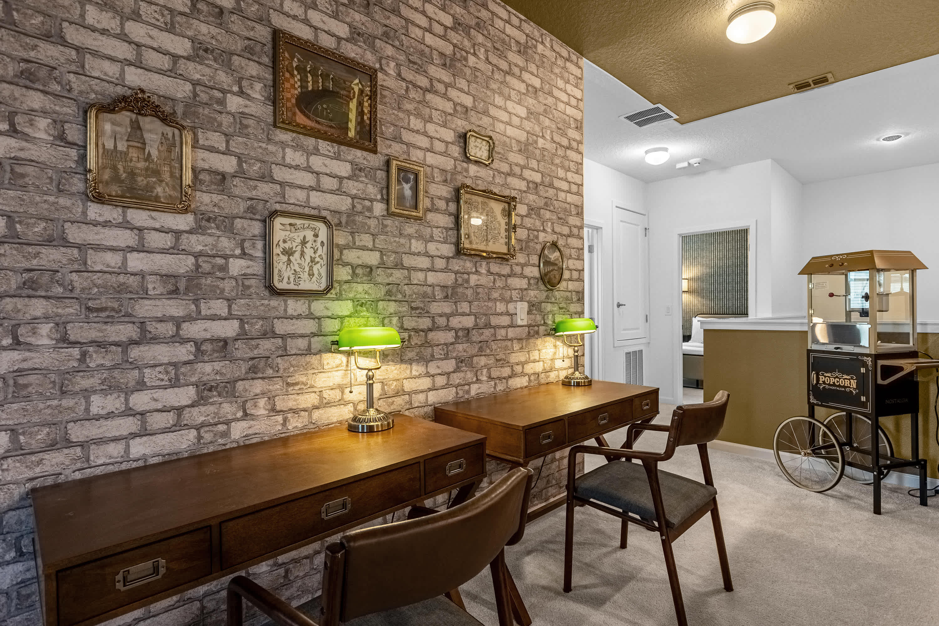 A charming study nook or hallway area with faux brick walls adorned with vintage-style framed art. Two wooden desks with green-shaded lamps and chairs are visible, alongside a classic popcorn machine.
