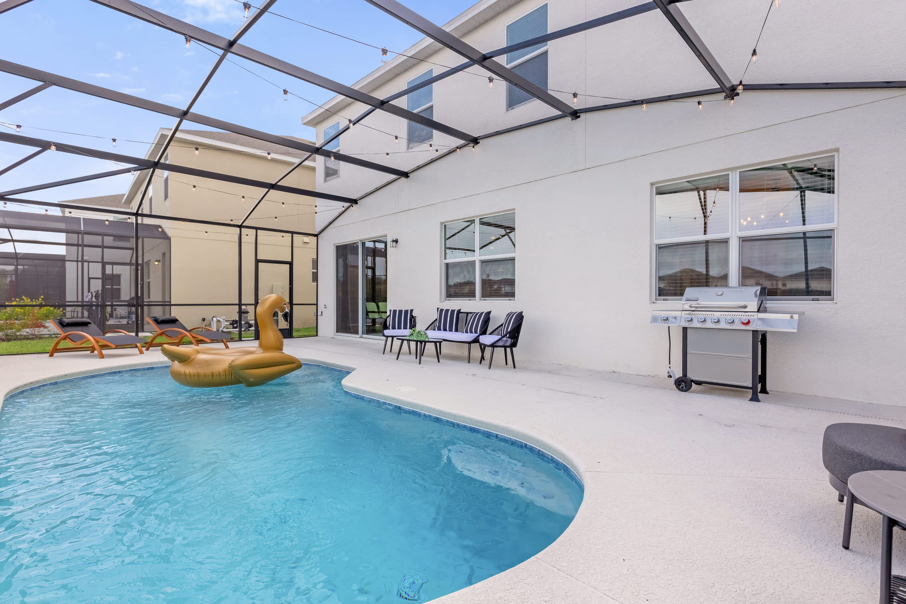 Screened-in private pool area with a freeform pool, a golden swan float, and a BBQ grill on the light concrete patio. Outdoor seating with striped cushions is visible, and string lights are strung across the screen enclosure.