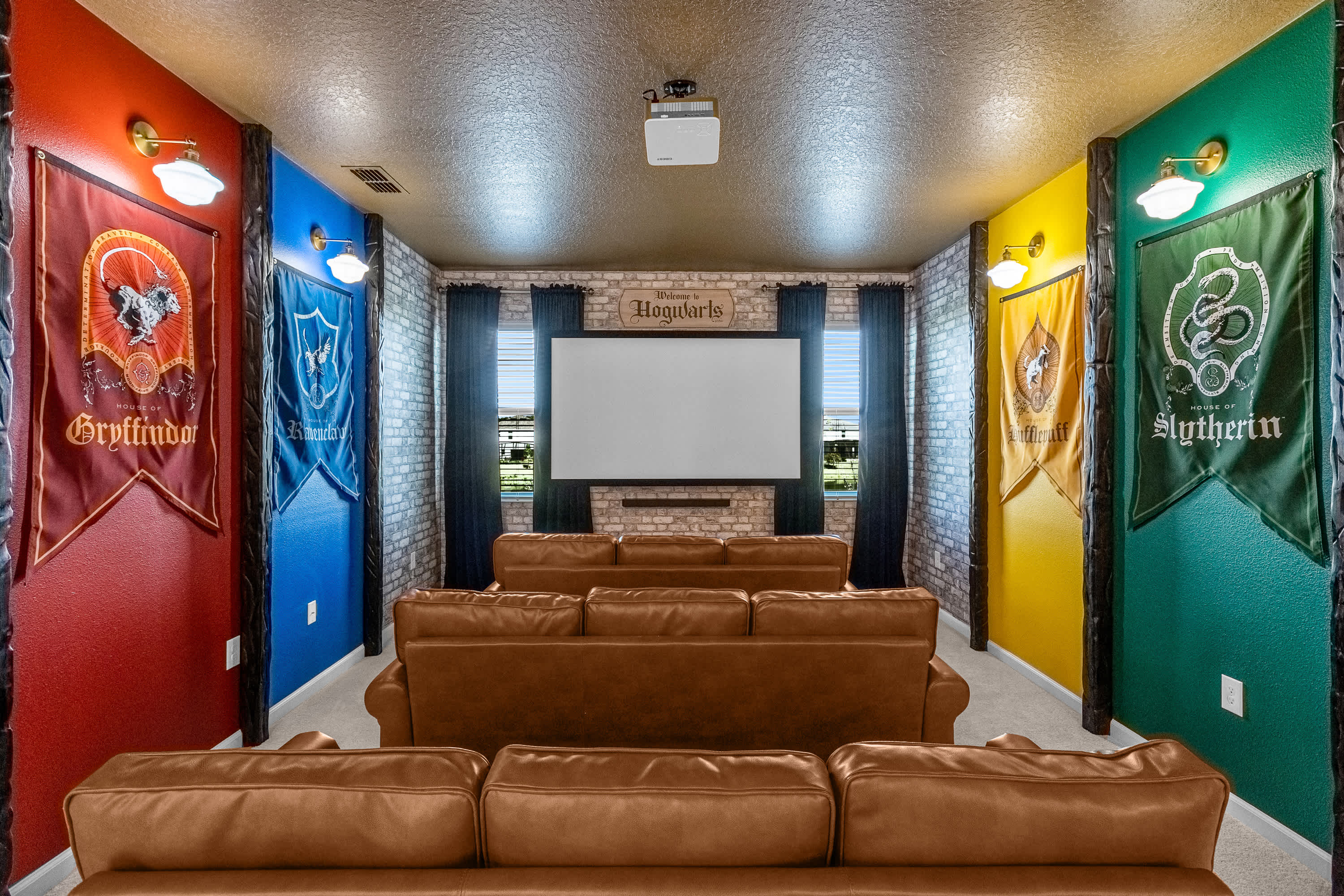 Harry Potter-themed home theater with rows of brown leather recliners facing a large projection screen. Walls are painted in house colors (red, blue, yellow, green) with corresponding banners, and brick-patterned wallpaper behind the screen.