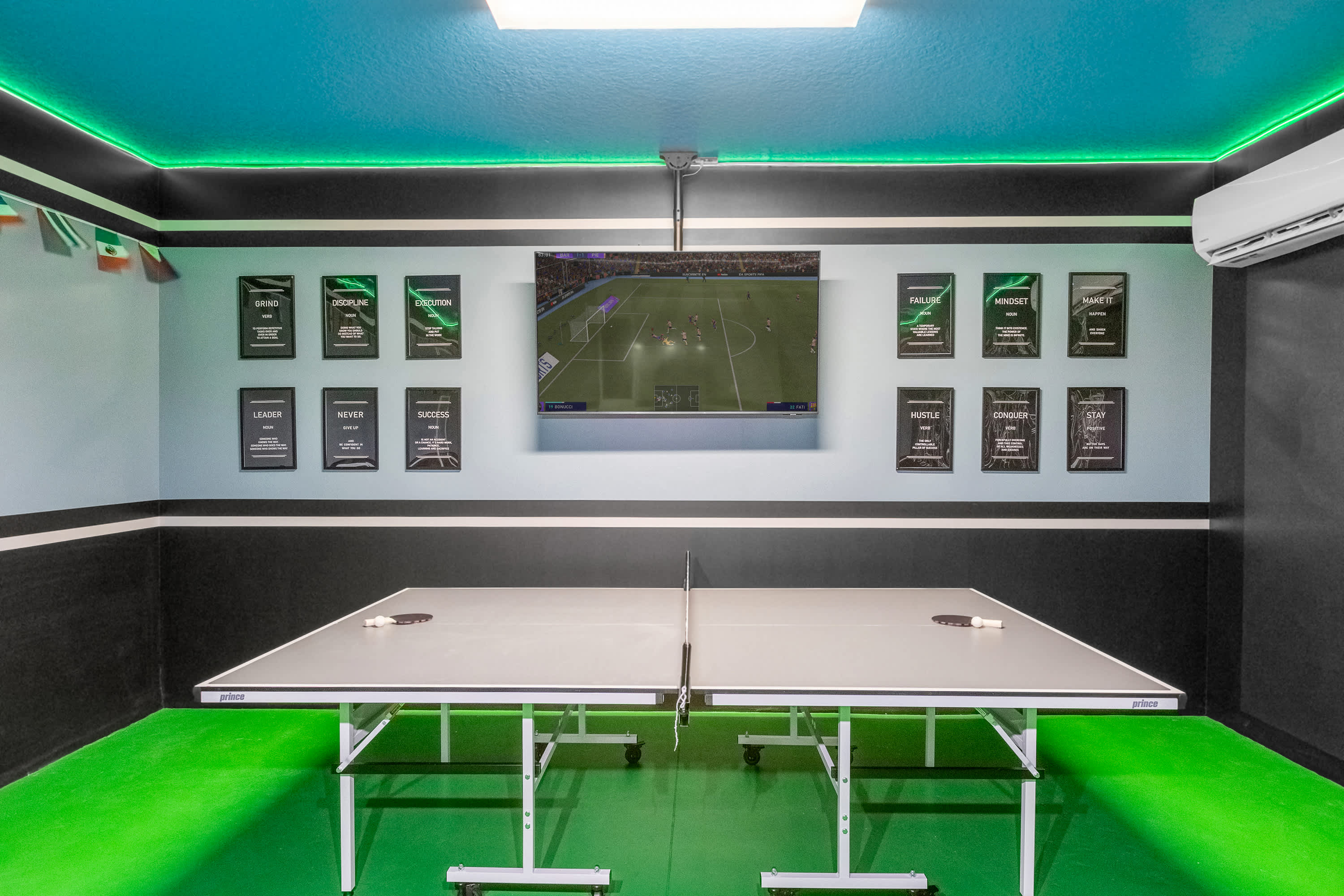 Game room with a vibrant green floor and ceiling, featuring two ping-pong tables. A large flat-screen TV is mounted on the wall, surrounded by framed motivational quotes, with green LED strip lighting along the ceiling.