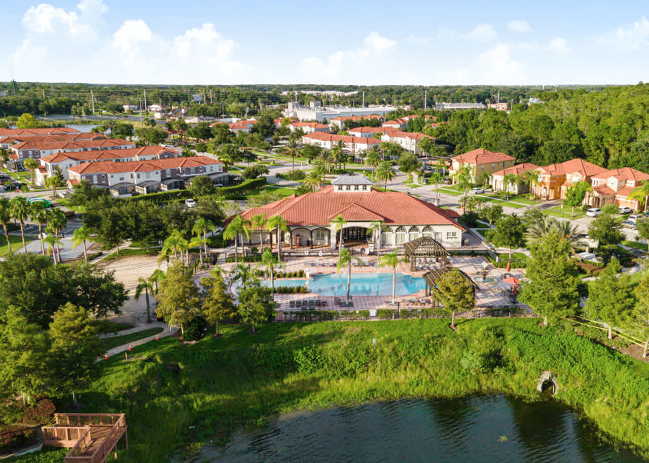 Wide aerial view of a residential community featuring a central clubhouse and large swimming pool complex, surrounded by numerous homes with red tile roofs, all nestled beside a tranquil body of water and lush green areas.