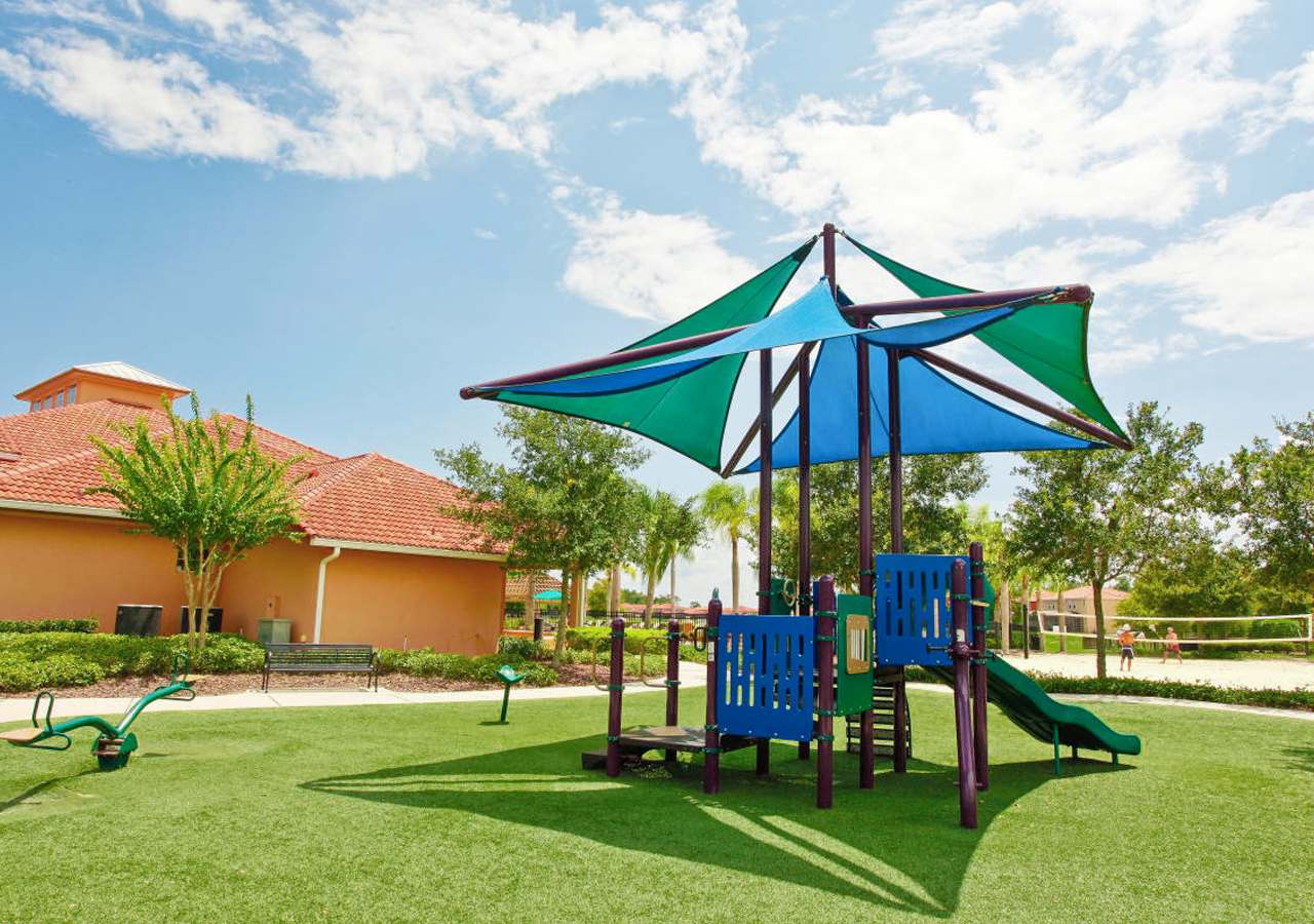 Vibrant community playground featuring a blue and green shade structure over a play set with a slide and climbing elements, alongside a green seesaw, all situated on lush artificial turf with a clubhouse building in the background.