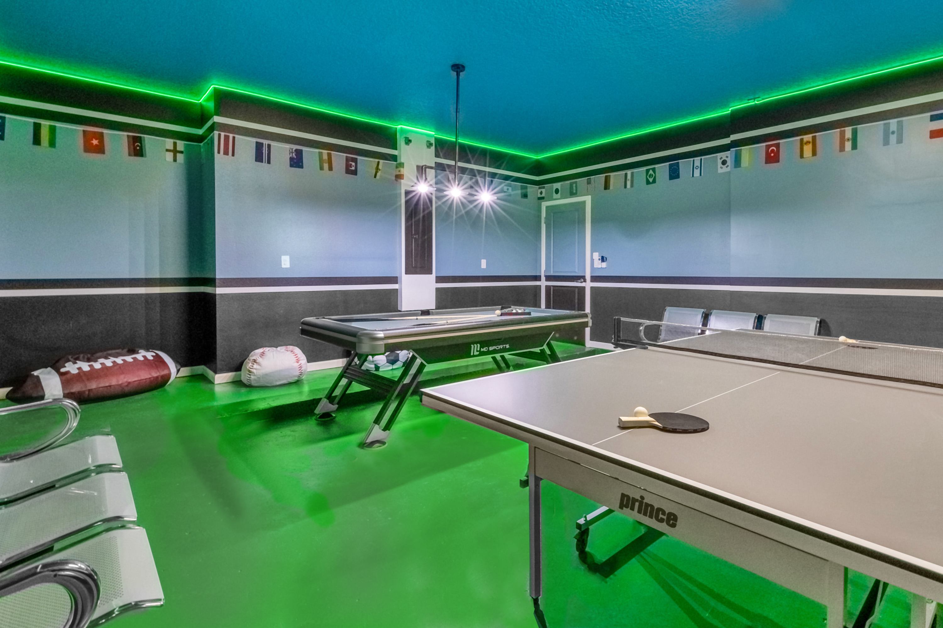 A vibrant game room with a bright green floor, blue ceiling, and neon green strip lighting. It features a pool table, a ping-pong table, and sports-themed decorations including international flags and a large football beanbag.