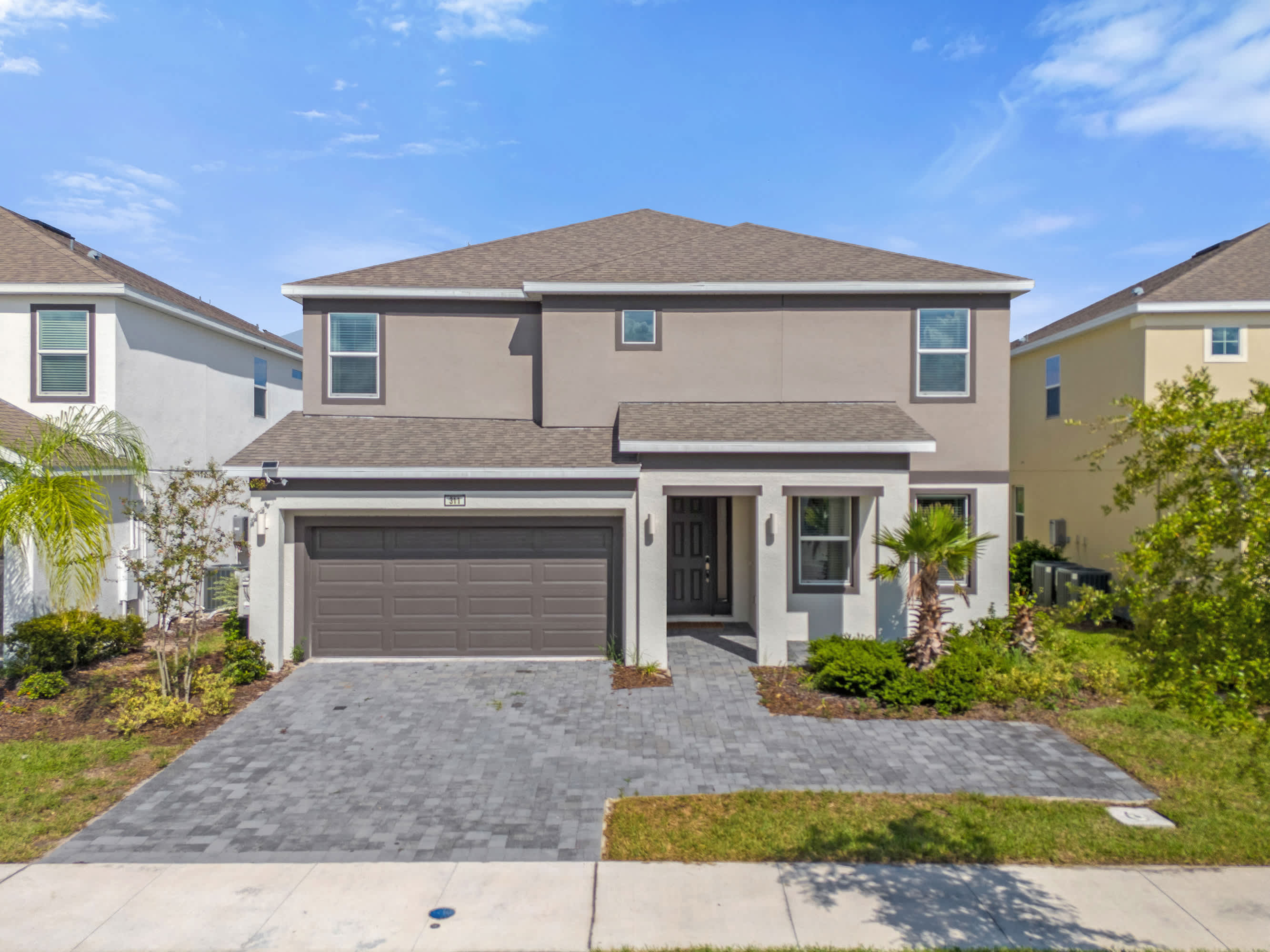 An inviting two-story modern house with a light gray stucco exterior, dark gray roof, and a paved driveway leading to a two-car garage, surrounded by manicured landscaping and palm trees.