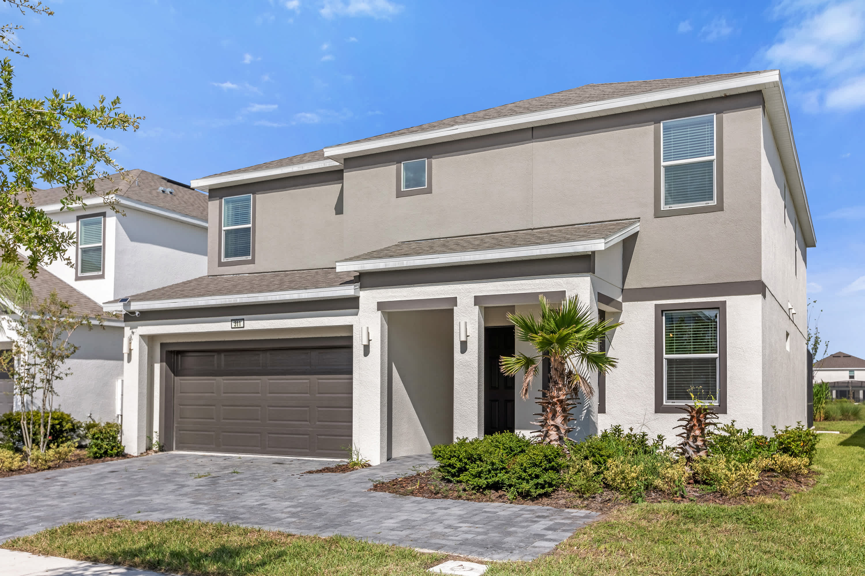 Contemporary two-story house with a light grey and white stucco exterior, a dark grey garage door, and a covered entryway with a palm tree. The front yard features low-maintenance landscaping and a paved driveway.