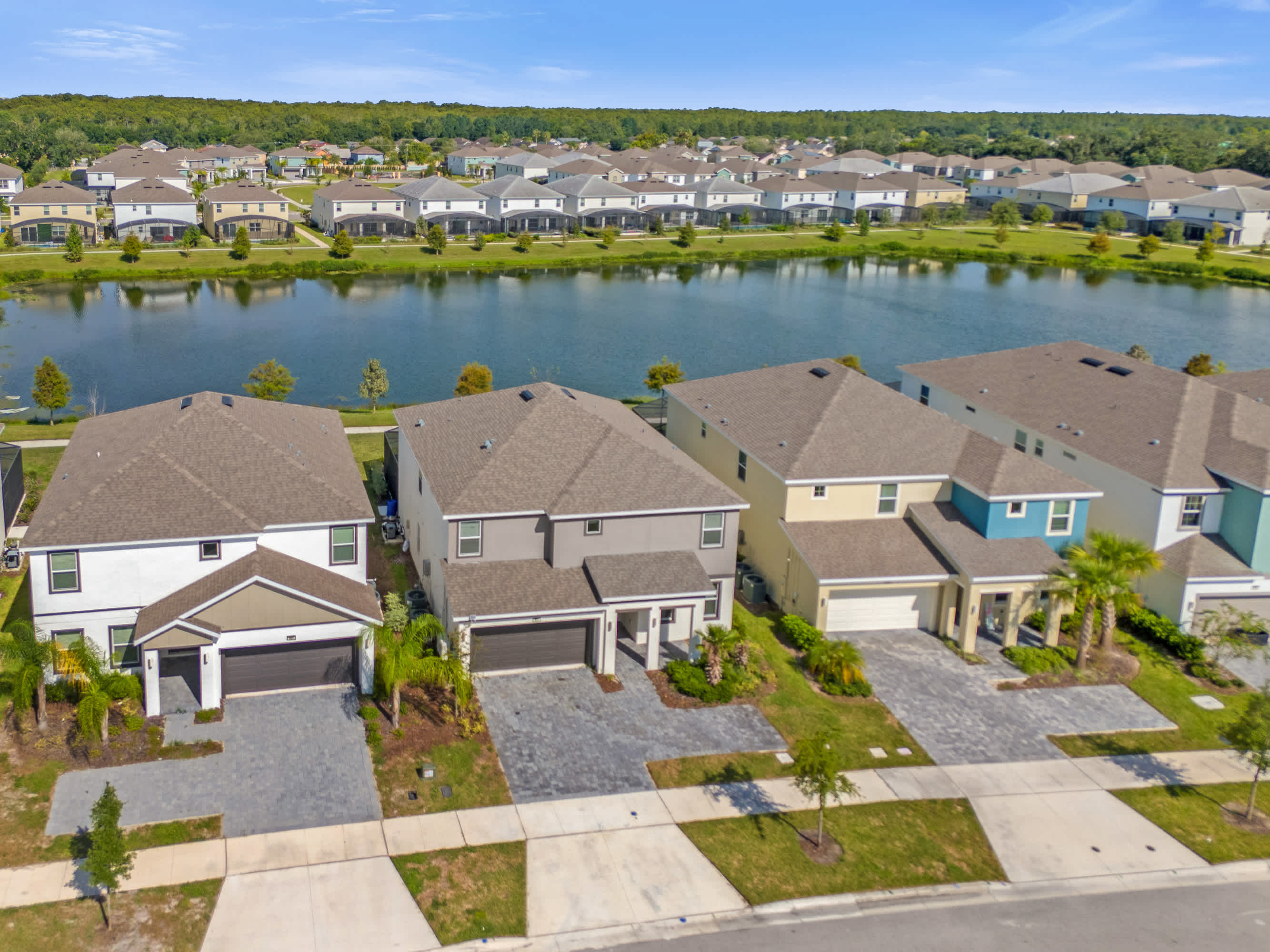 An aerial view of a residential community featuring rows of modern houses with varying exterior colors, situated along the edge of a serene lake under a clear blue sky.