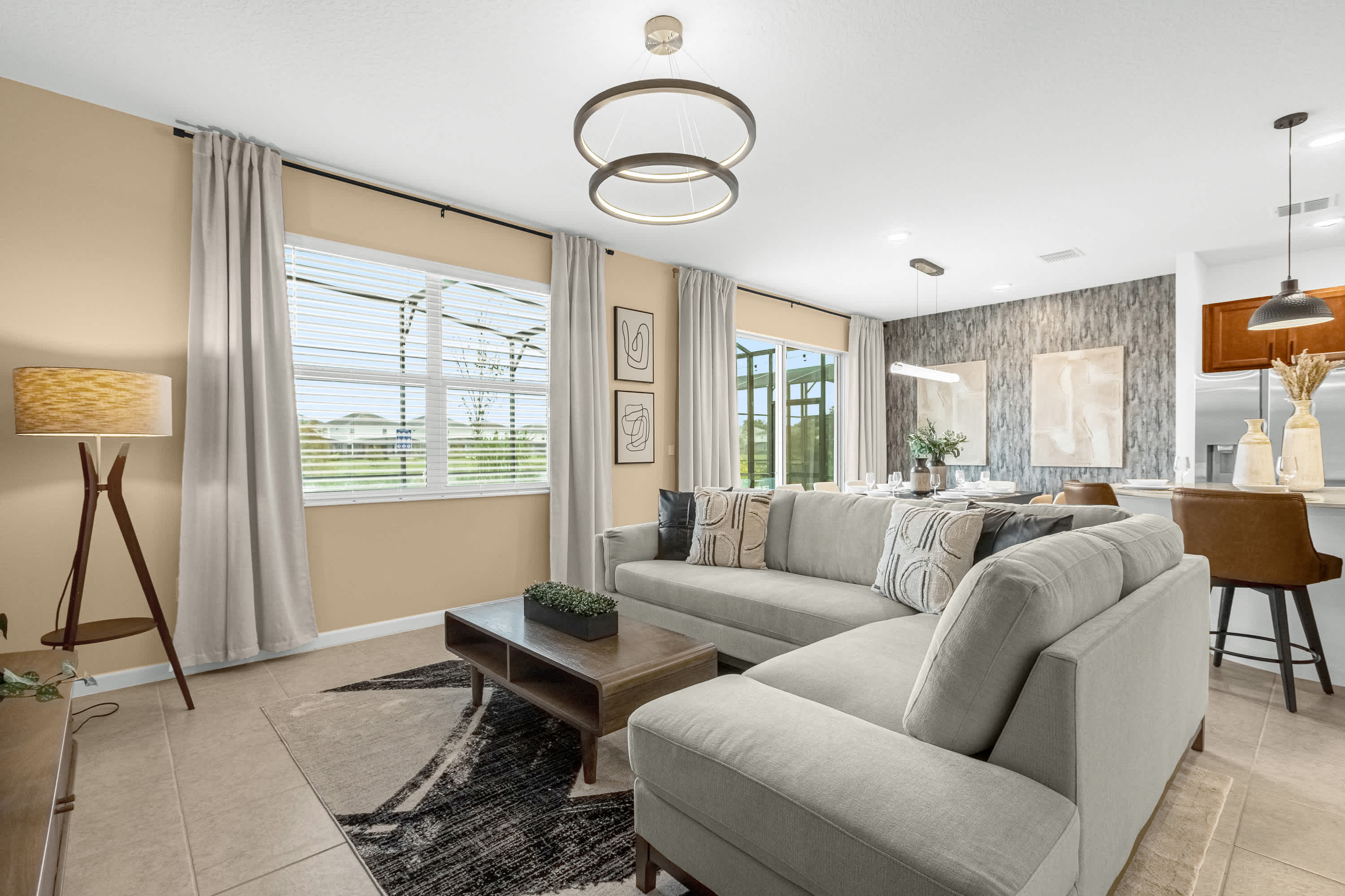 Bright living room with a large light grey sectional sofa and a dark wood coffee table on a patterned rug. The room features light beige walls, modern circular chandeliers, and views of the screened patio and pool area through large windows.