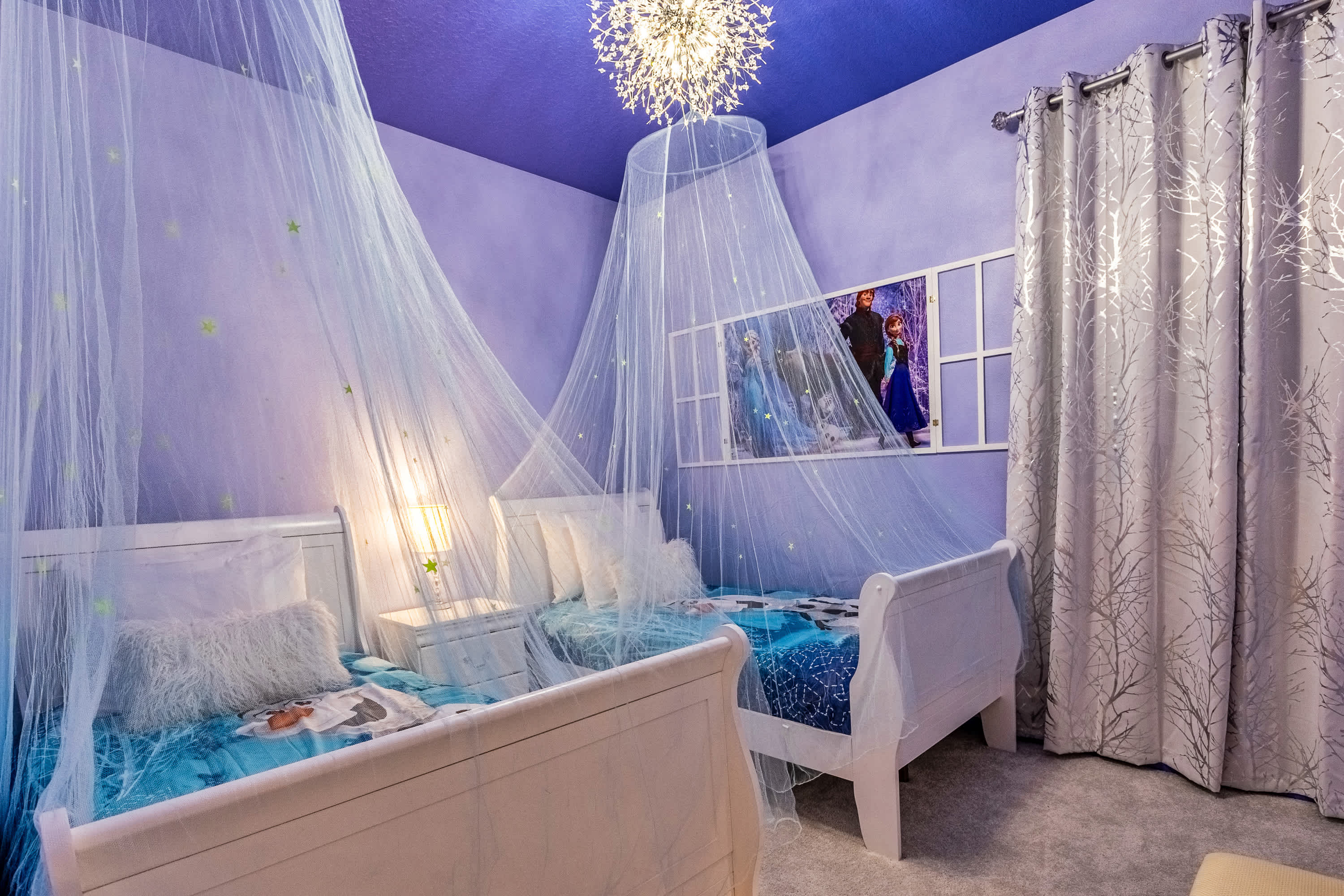 A themed children's bedroom with two twin beds, each featuring a sheer canopy and themed bedding. The room has lavender walls and ceiling, a sparkling chandelier, and a large framed artwork depicting characters from Frozen above the beds.