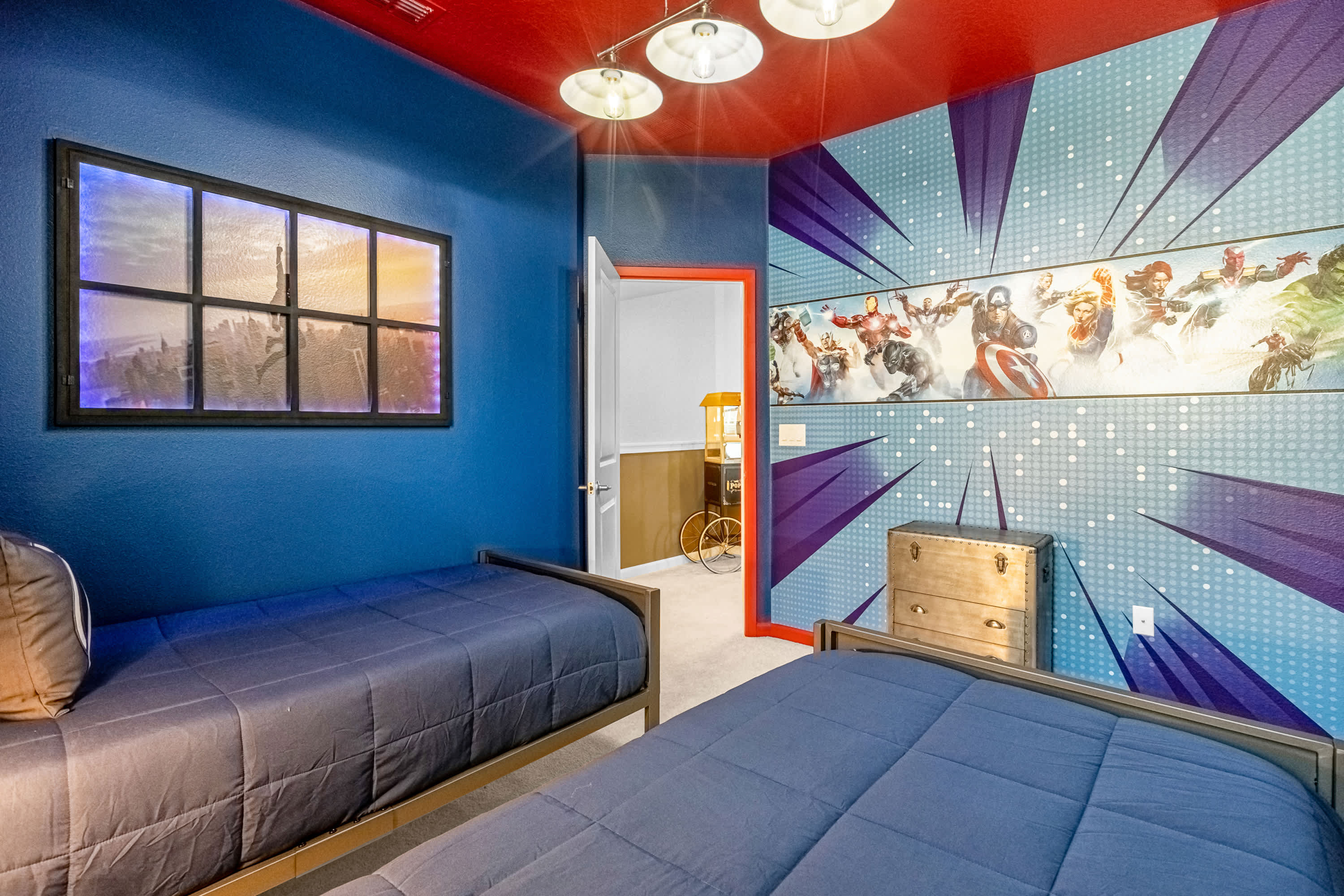 Vibrant superhero-themed bedroom with two twin beds, bright blue walls, and a red ceiling. A large mural of superheroes dominates one wall, and a unique window-like light fixture adds to the theme.