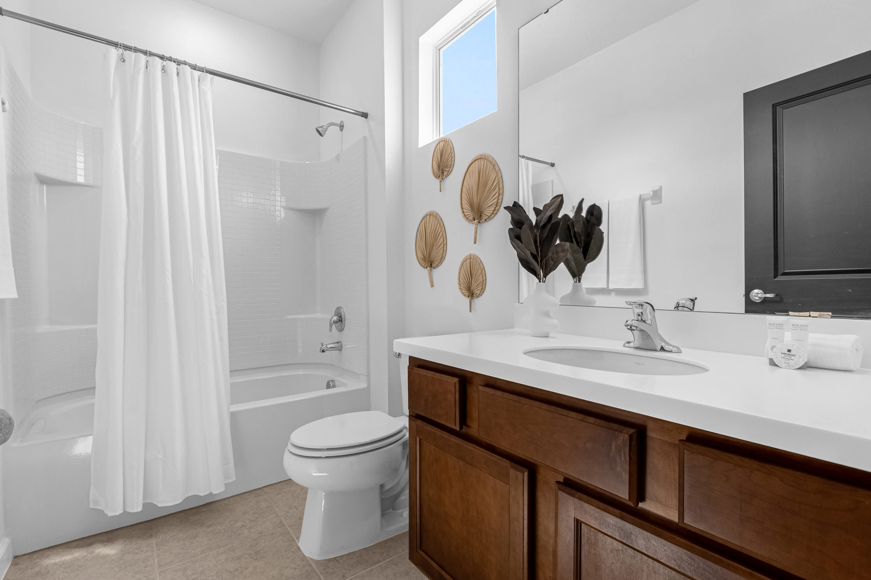 Bright bathroom with a single vanity featuring a white countertop and dark wood cabinets, a toilet, and a shower/tub combo with a white curtain. Decorative dried palm leaves adorn the wall.