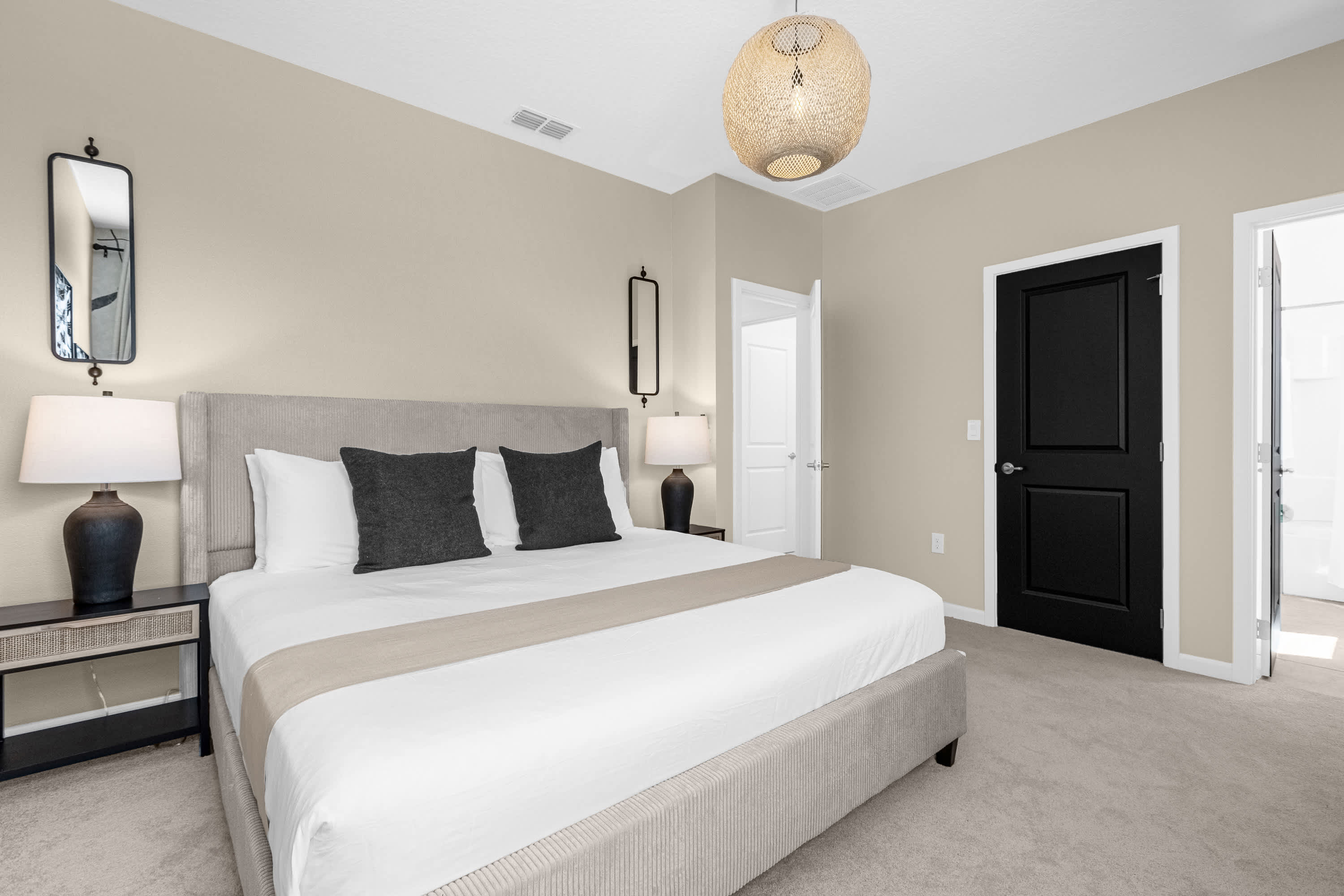 Spacious bedroom featuring a king bed with a tall upholstered headboard, two nightstands with lamps, and a large mirror. The walls are light beige, and a black door adds a modern contrast.