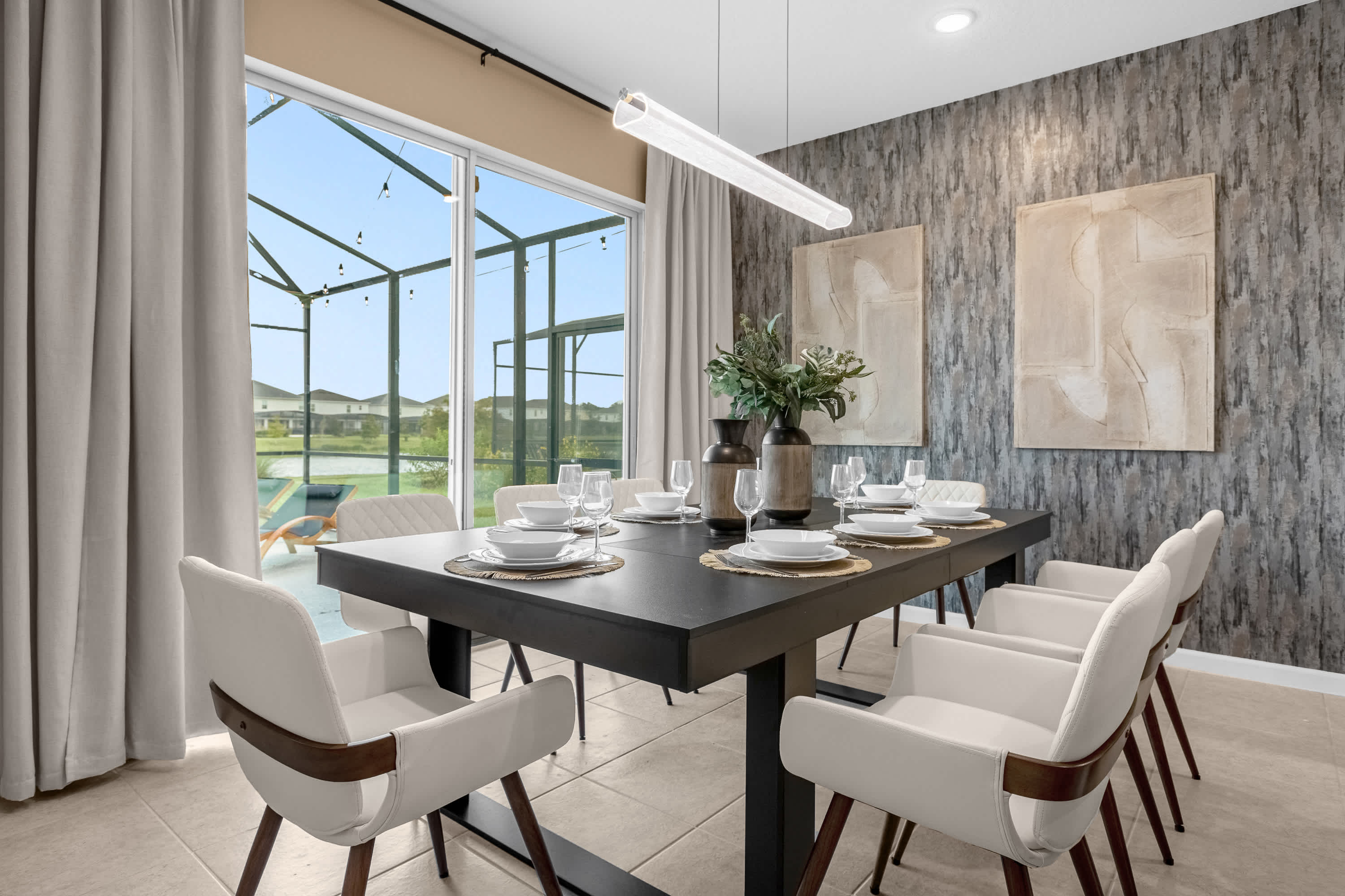 Modern dining room with a long black table set for eight, white upholstered chairs, and a linear pendant light. A large glass sliding door opens to an outdoor screened patio with string lights and a pool view.