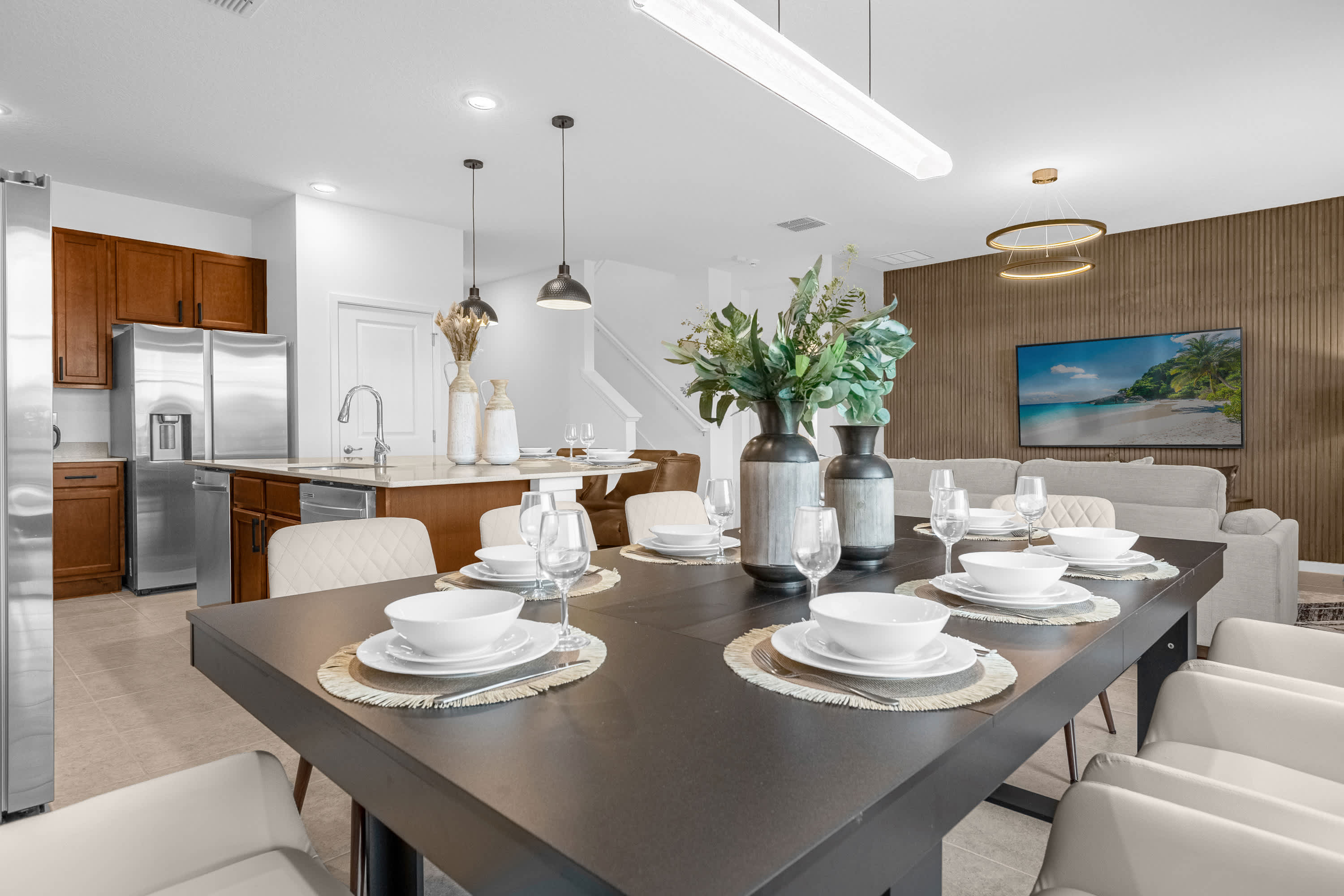 Open-concept dining area with a black table and white chairs, flowing into a living room with a large TV mounted on a textured accent wall. The space features modern lighting and views of the outdoor patio.