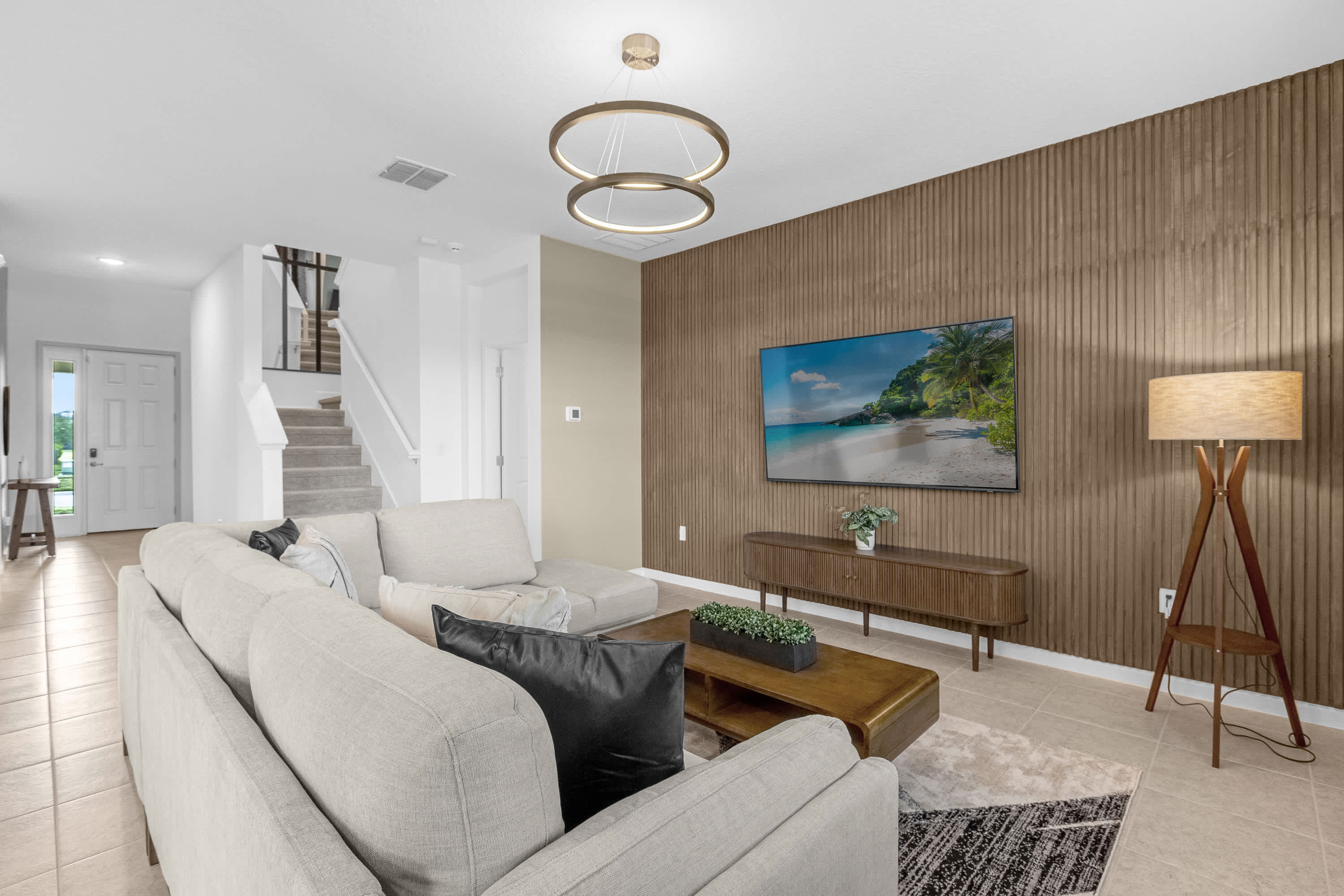 Cozy living room with a large light-colored sectional sofa and a wooden media console beneath a wall-mounted TV. A textured wooden accent wall adds warmth, and a modern floor lamp stands beside the sofa.