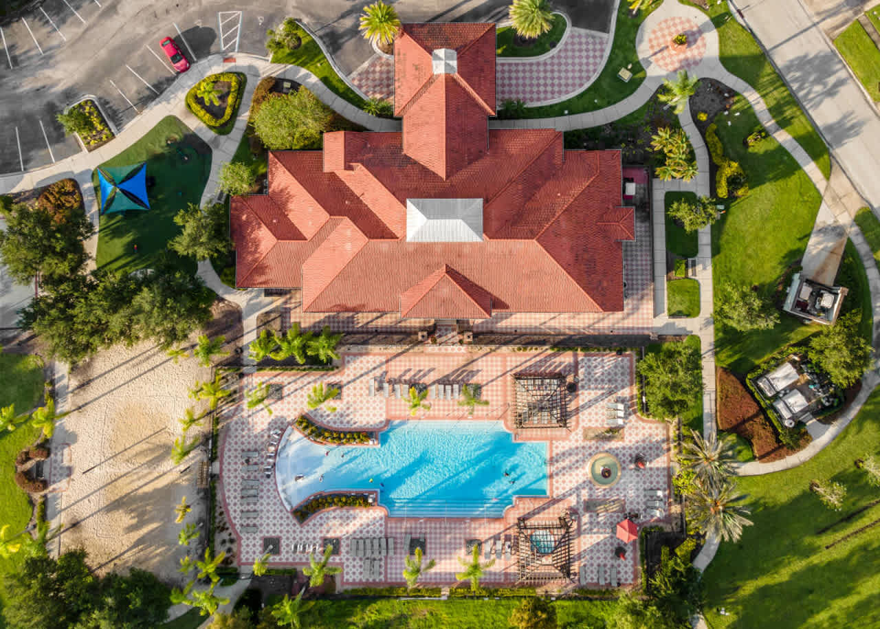 Overhead aerial view showcasing a large community clubhouse with a red tile roof, an expansive swimming pool with sun loungers, multiple pergolas, a decorative fountain, and a sand volleyball court, surrounded by manicured landscaping.
