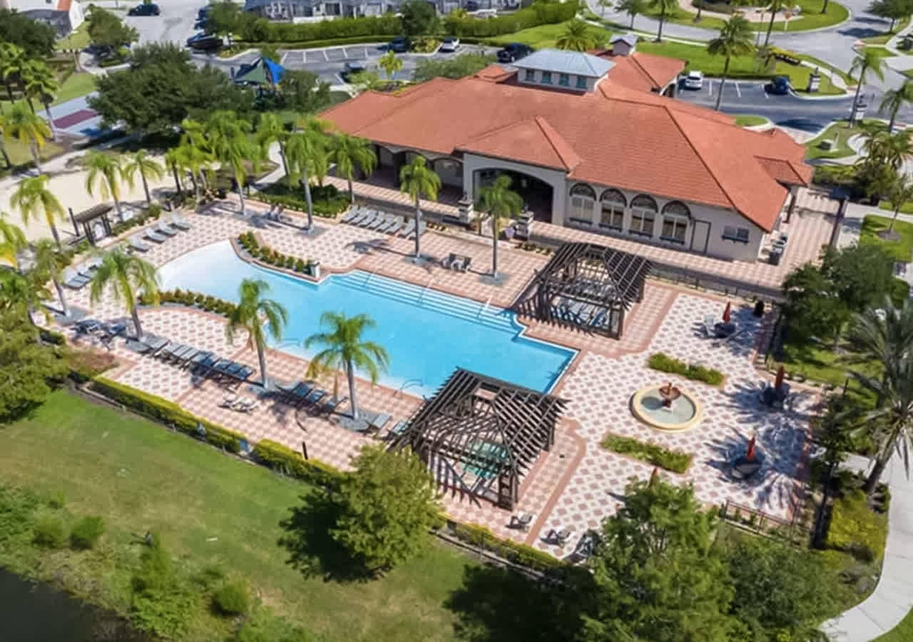 Aerial view of a large community clubhouse with a red tile roof, featuring a spacious outdoor swimming pool surrounded by numerous lounge chairs, paved sun decks, and shaded pergolas, all set amidst lush green landscaping and palm trees.