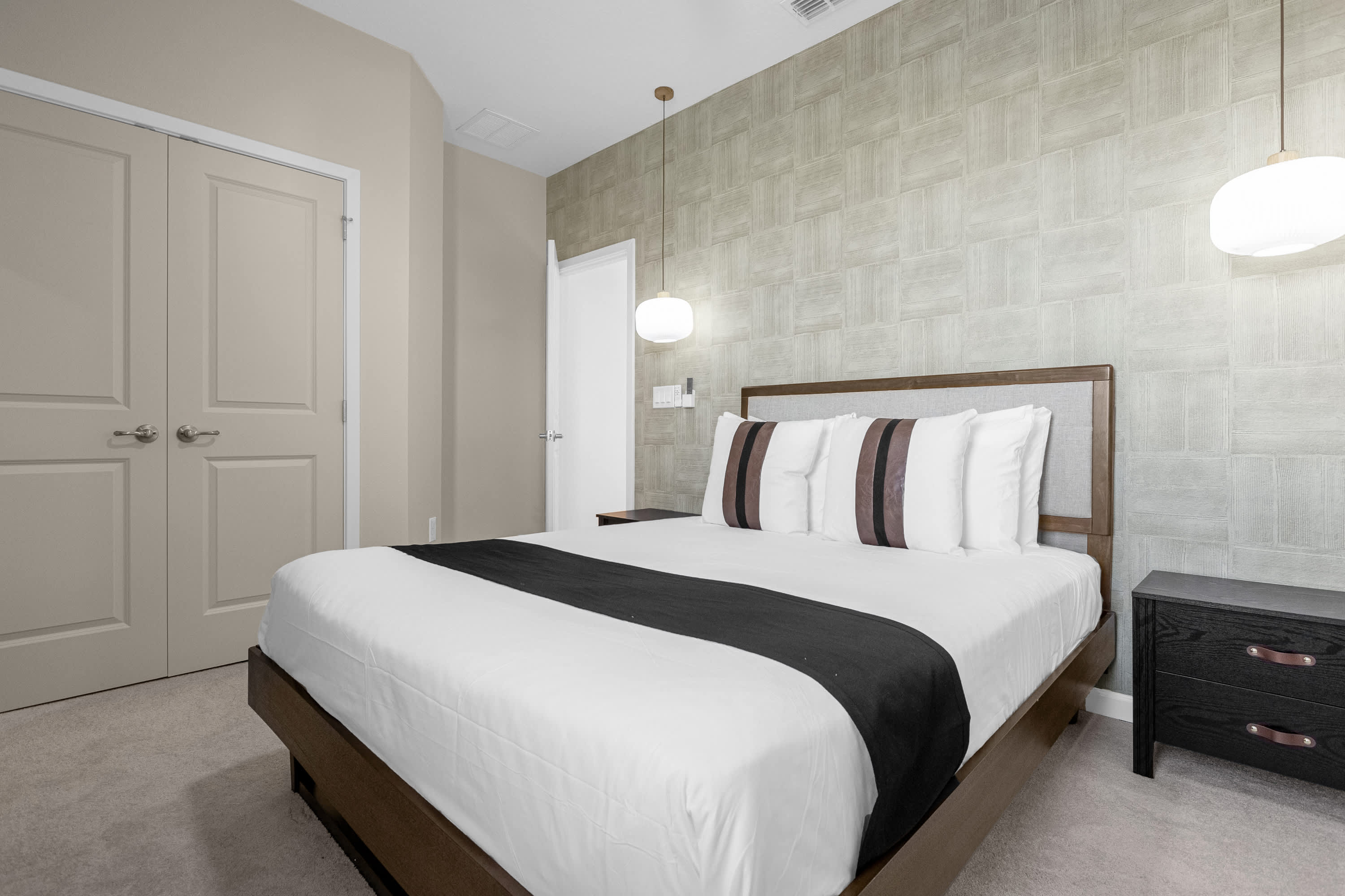Elegant bedroom featuring a queen bed with a wooden headboard, two small nightstands with pendant lights, and a textured light green accent wall. Two light-colored closet doors are visible.