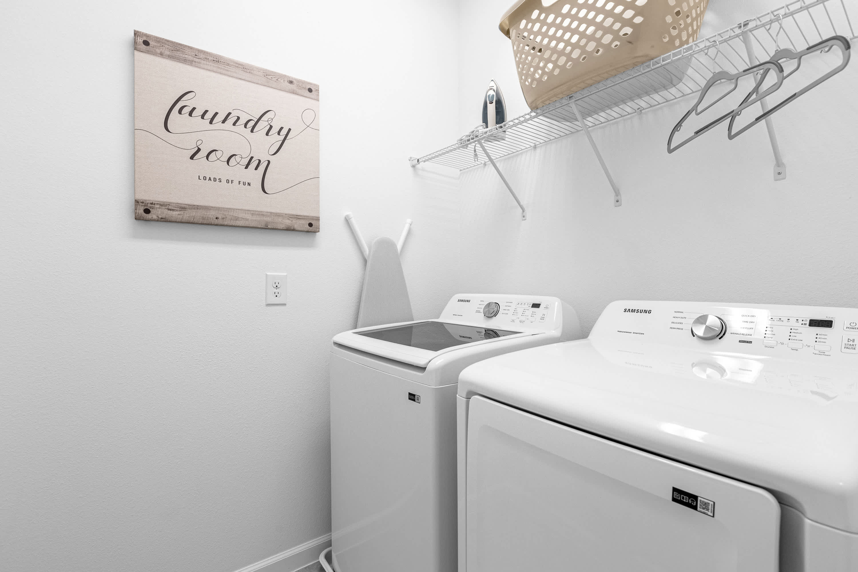 A functional laundry room equipped with a white top-load washing machine and a front-load dryer, a wall-mounted shelf for storage, and a decorative "laundry room" sign.