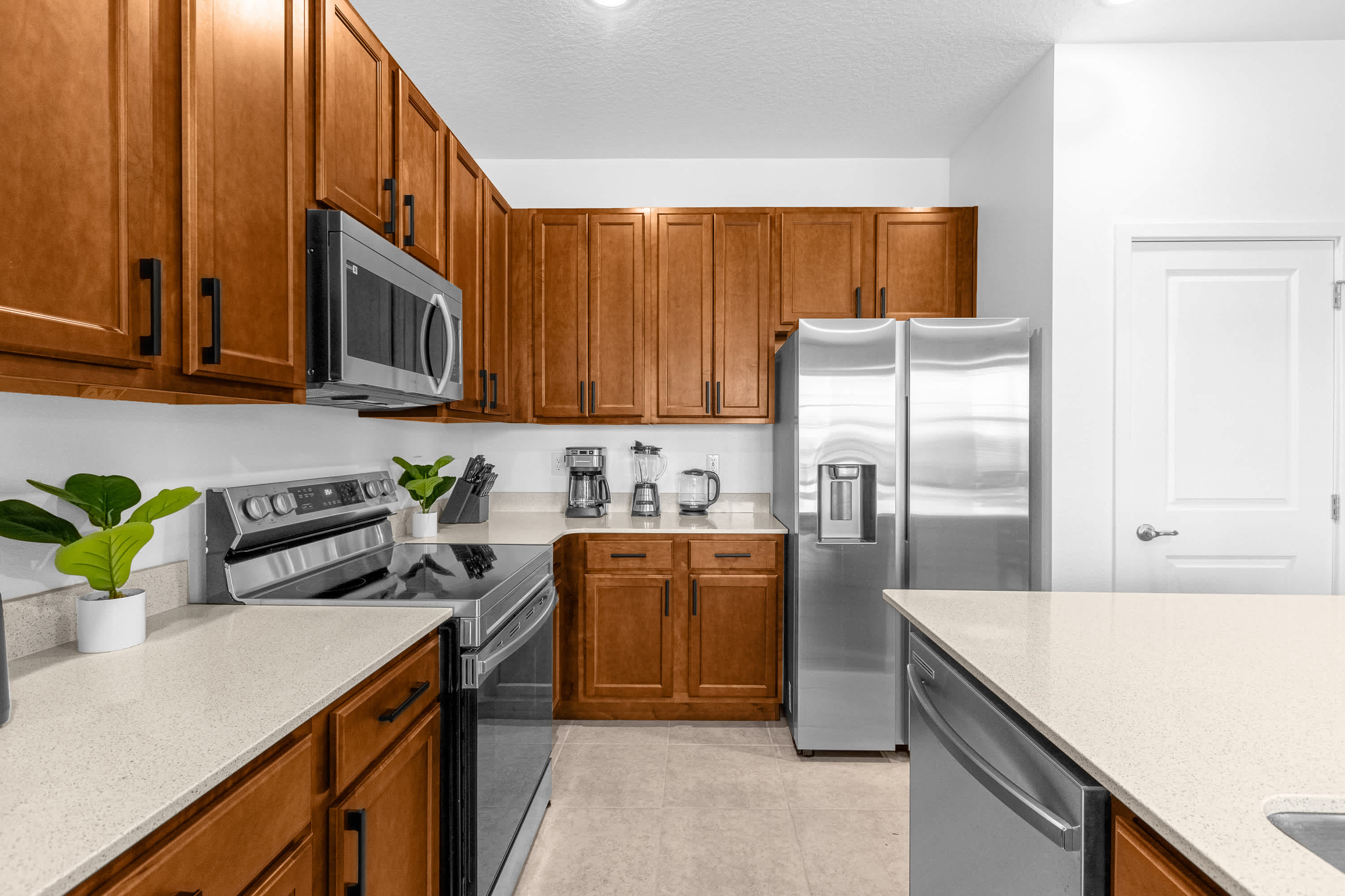Well-equipped kitchen with warm wooden cabinetry, light quartz countertops, and stainless steel appliances including a microwave, range, and side-by-side refrigerator. A white pantry door is visible on the right.