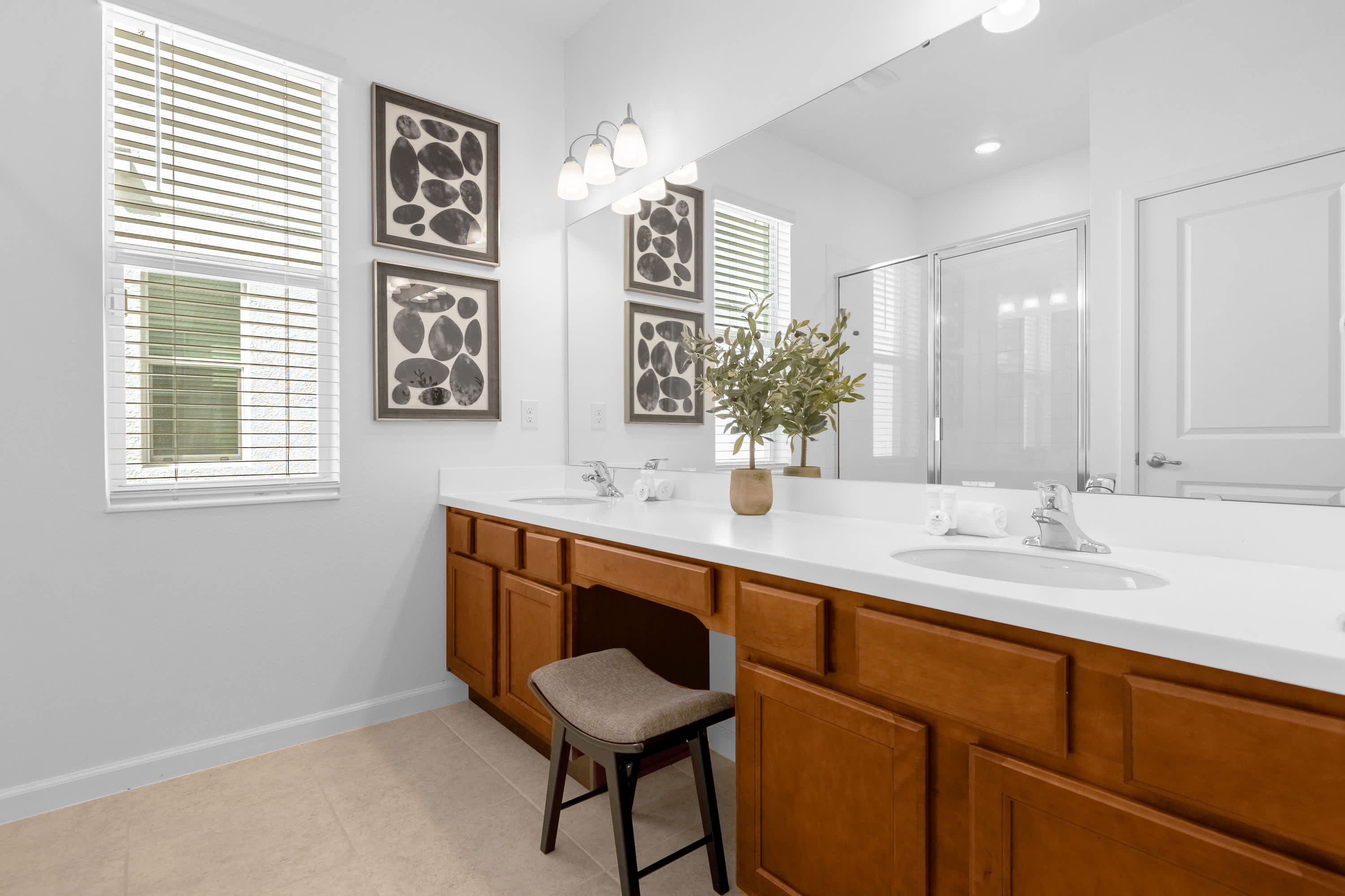 A spacious double vanity bathroom with light wood cabinets, white countertops, and two sinks. A large mirror spans the length of the vanity, which also includes a makeup vanity area with a stool, and two windows with blinds.