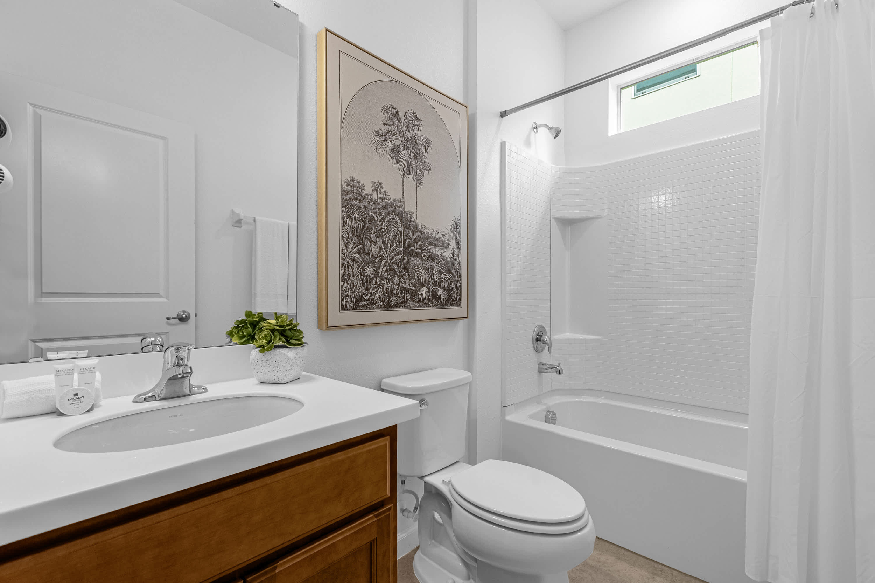 A bright white bathroom featuring a single vanity with a light wood cabinet and white countertop, alongside a toilet and a bathtub with a shower. The walls are adorned with white tiles, and a small window is positioned above the tub.