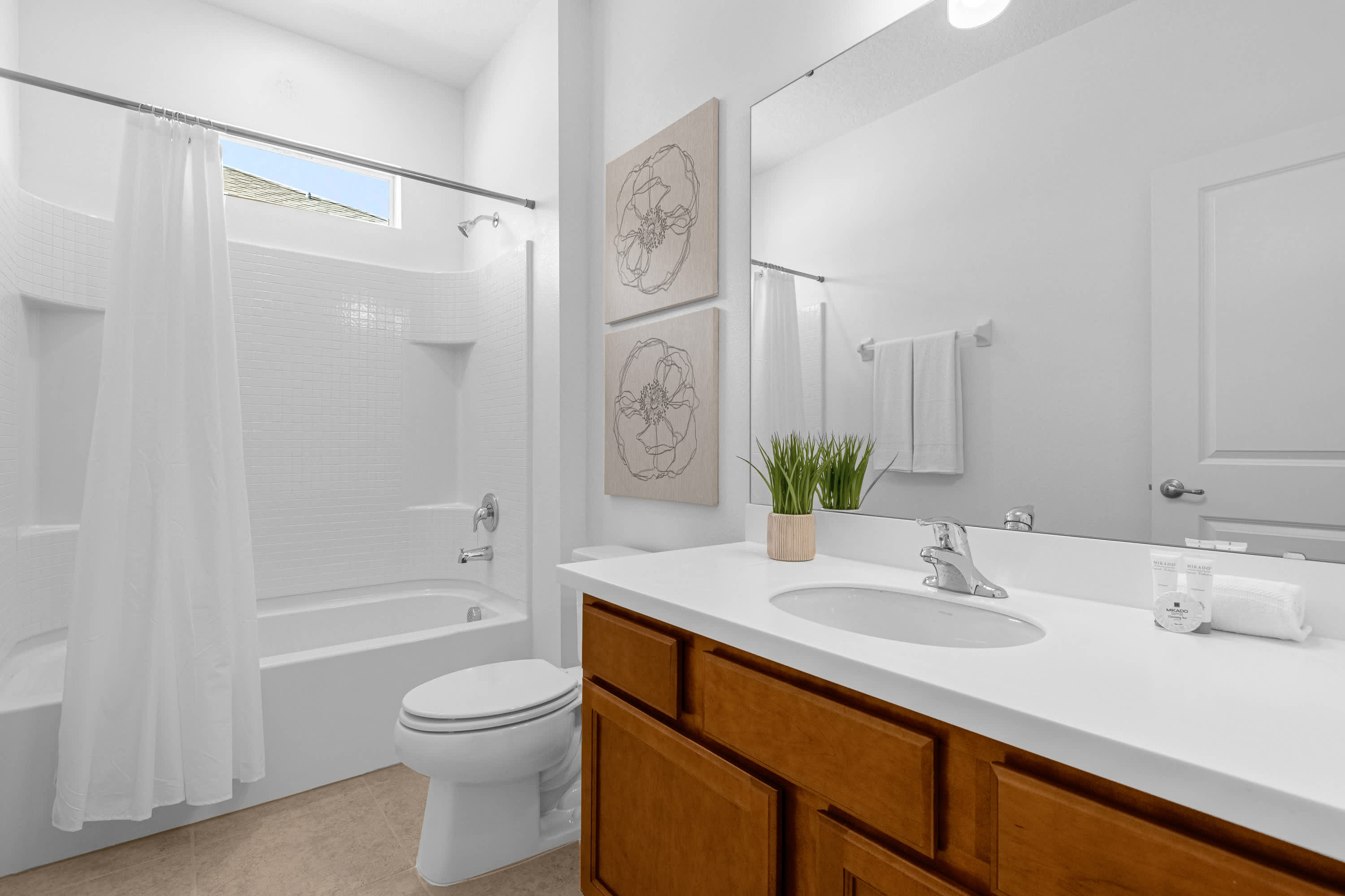 A bright bathroom with a single vanity featuring a light wood cabinet and white countertop, adorned with a small potted plant. It includes a toilet and a bathtub with a shower, all set against white tiled walls and a window above the tub.