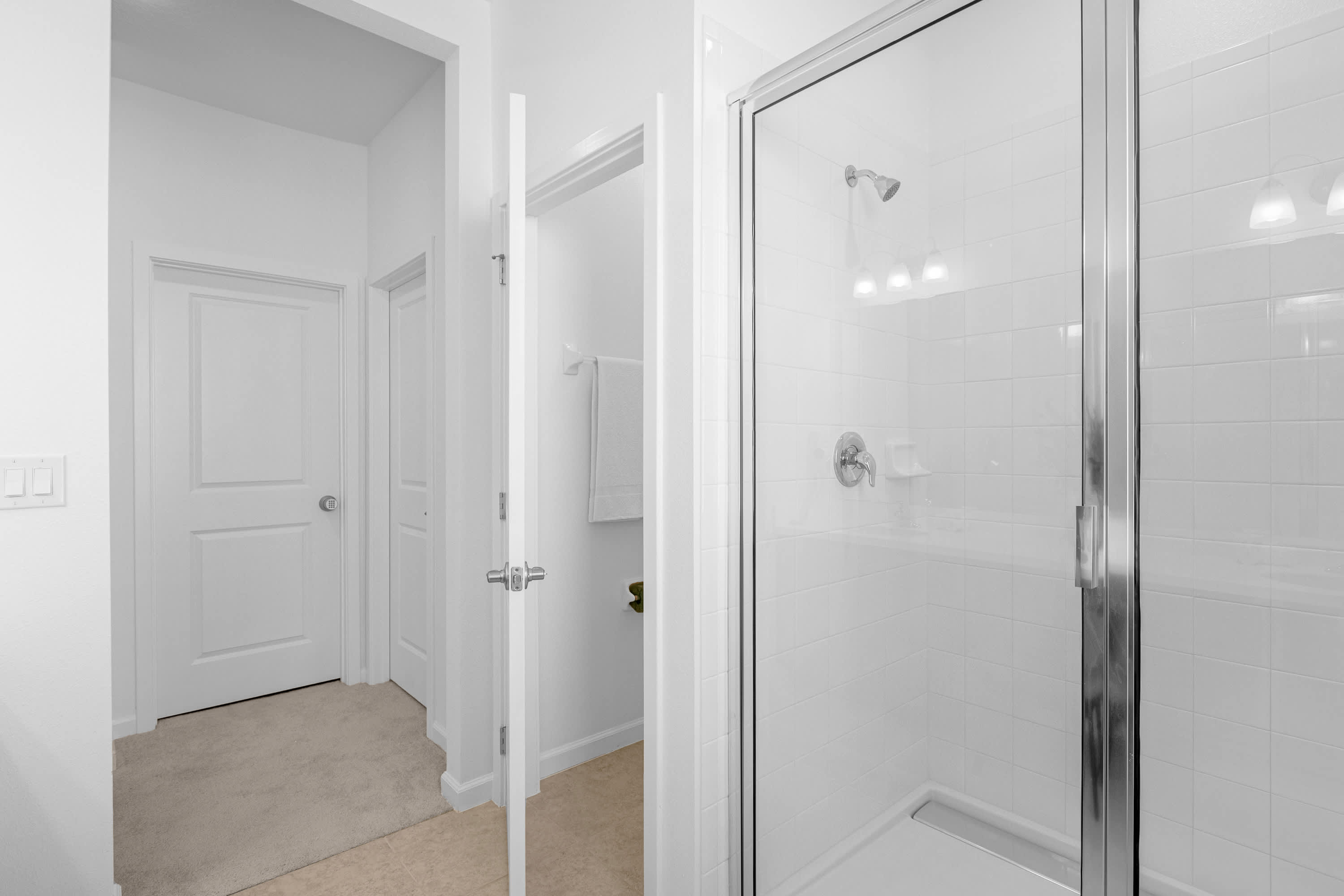 A bright, minimalist bathroom featuring a walk-in shower with white subway tiles and a clear glass enclosure. The room has light-colored floor tiles and leads to a separate toilet area, maintaining a clean and modern aesthetic.