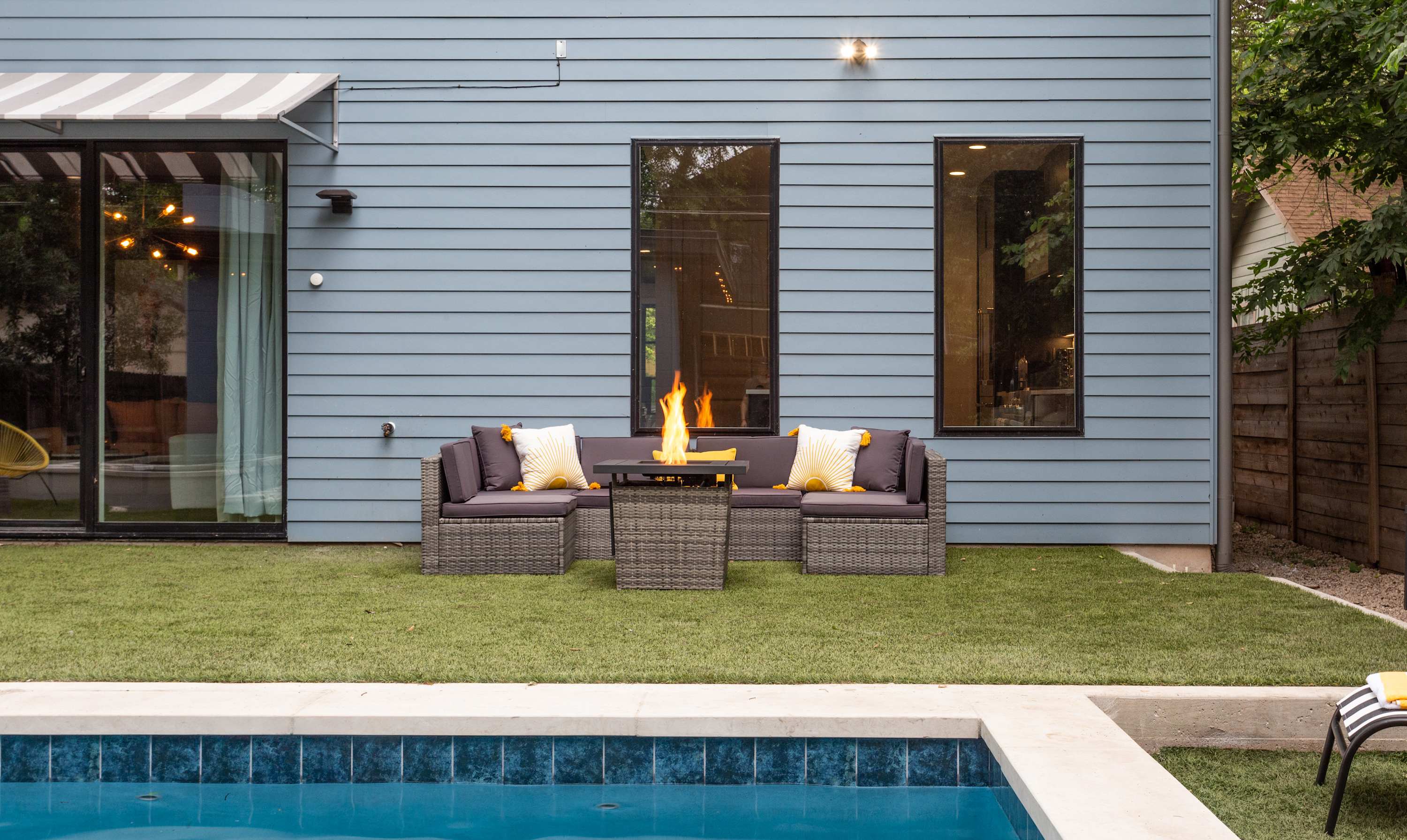 A view of the backyard from the pool area, showing a patio with a fire pit table and wicker outdoor seating. The house exterior features light blue siding and large windows, with artificial turf in the foreground.
