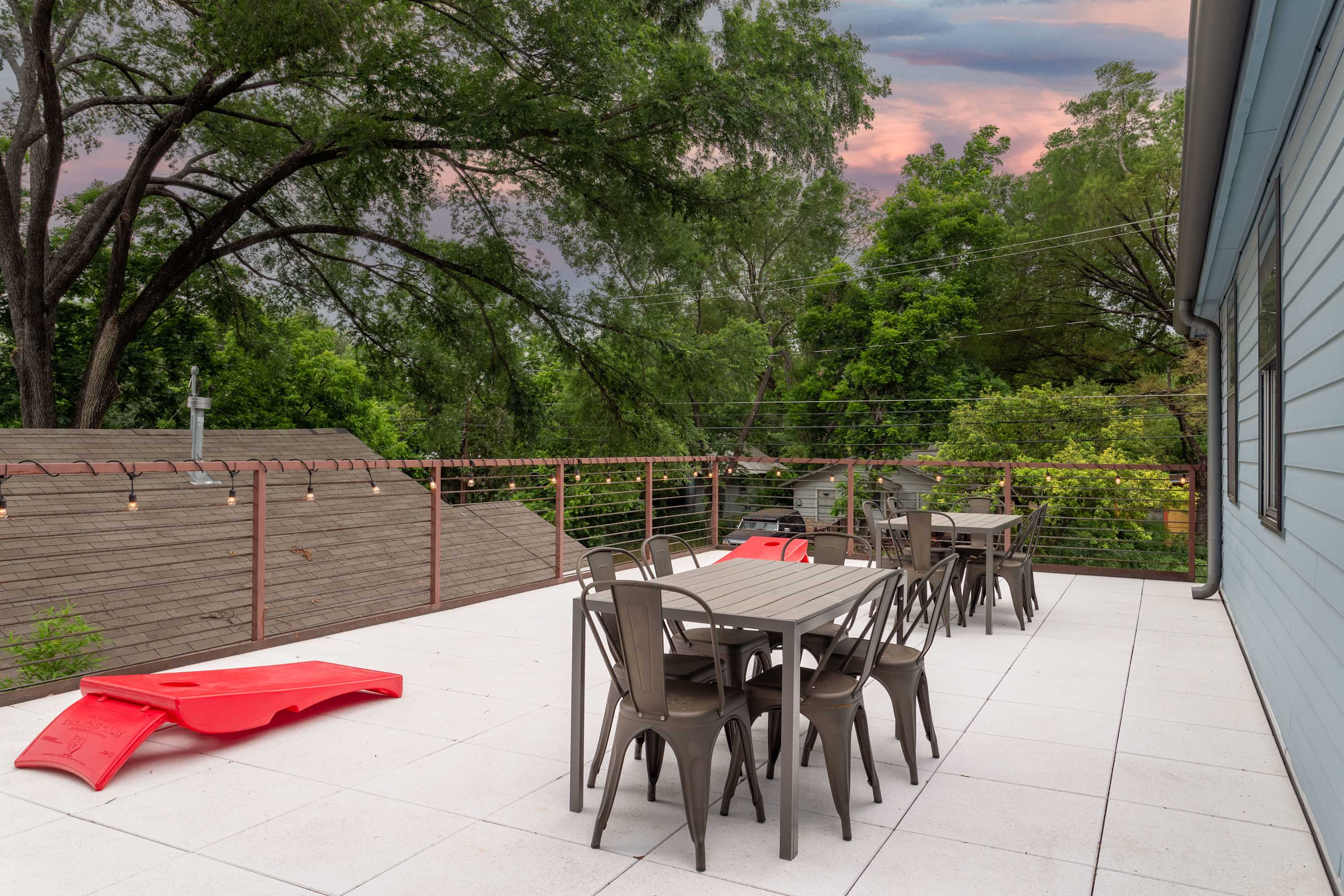 A spacious rooftop deck features multiple dark metal dining sets, string lights, and a red plastic slide, all set against a backdrop of lush green trees and a dramatic sunset sky.
