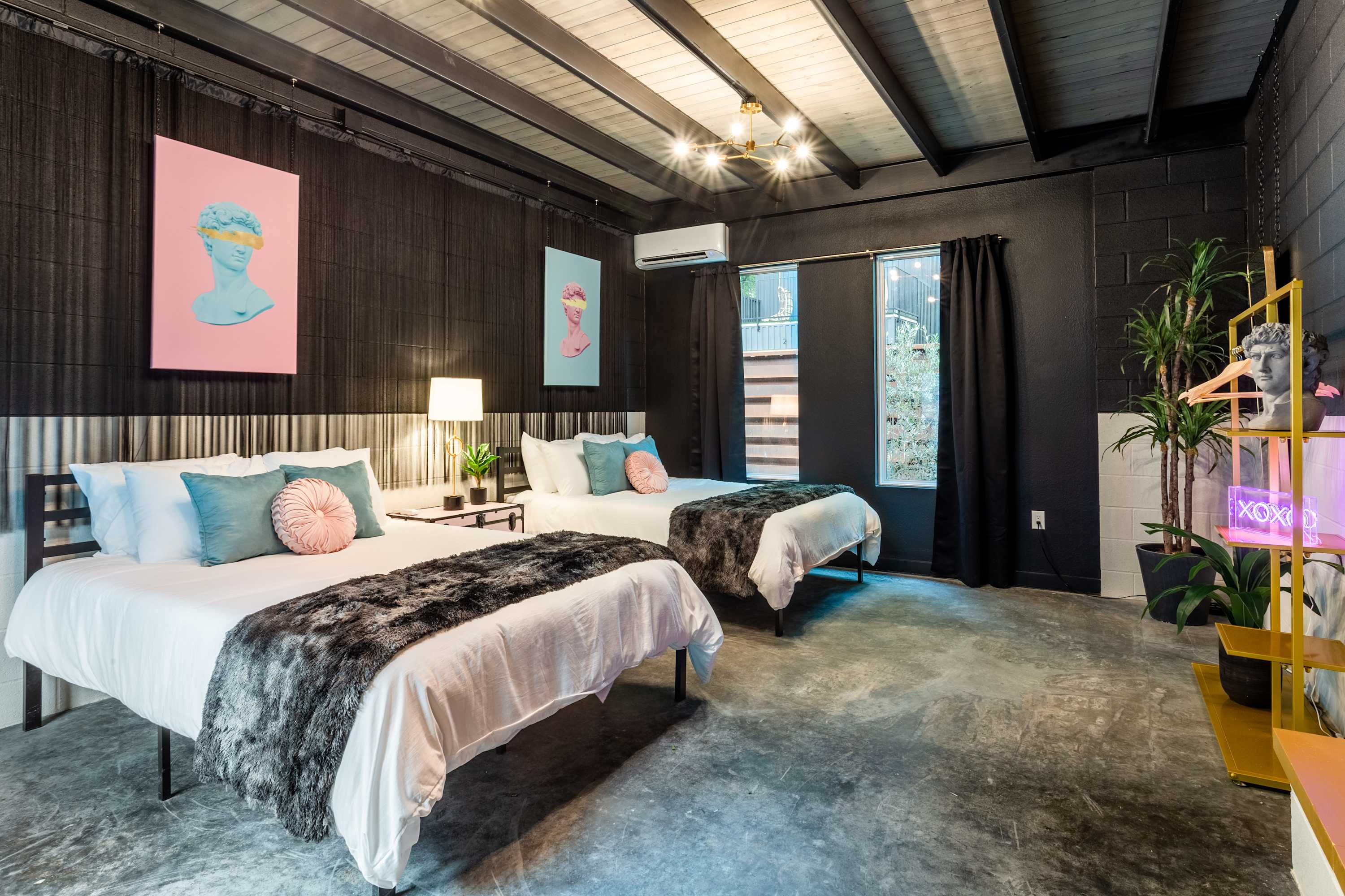 A modern bedroom featuring two queen beds with white bedding and plush faux fur throws, set against a textured black wall. The room has polished concrete floors, a black exposed beam ceiling, and vibrant pop art prints adding a splash of color.