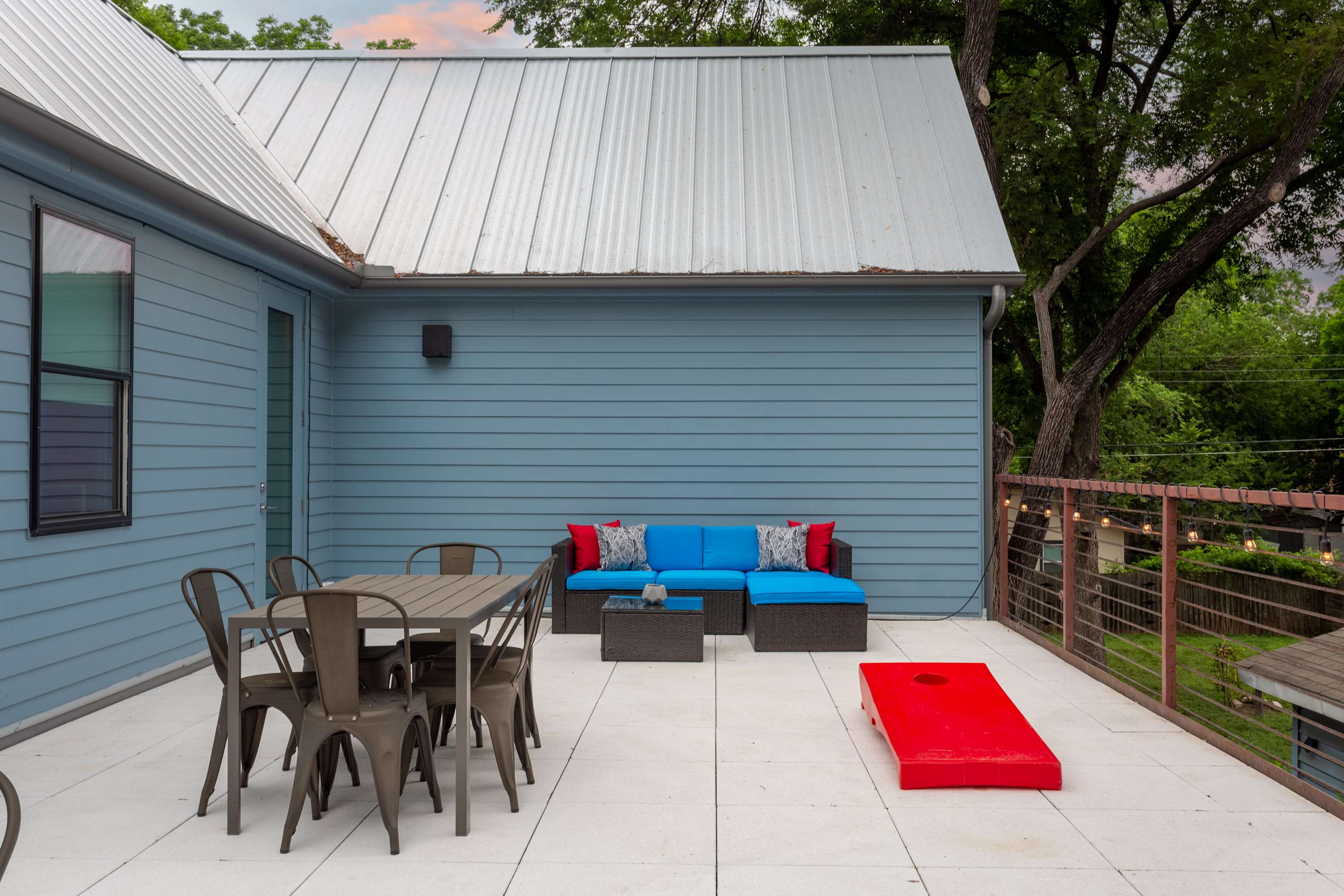 A spacious outdoor deck with a metal roof, featuring a dining table with chairs, a blue sectional sofa with red pillows, and a red cornhole game set. The house exterior has light blue siding.