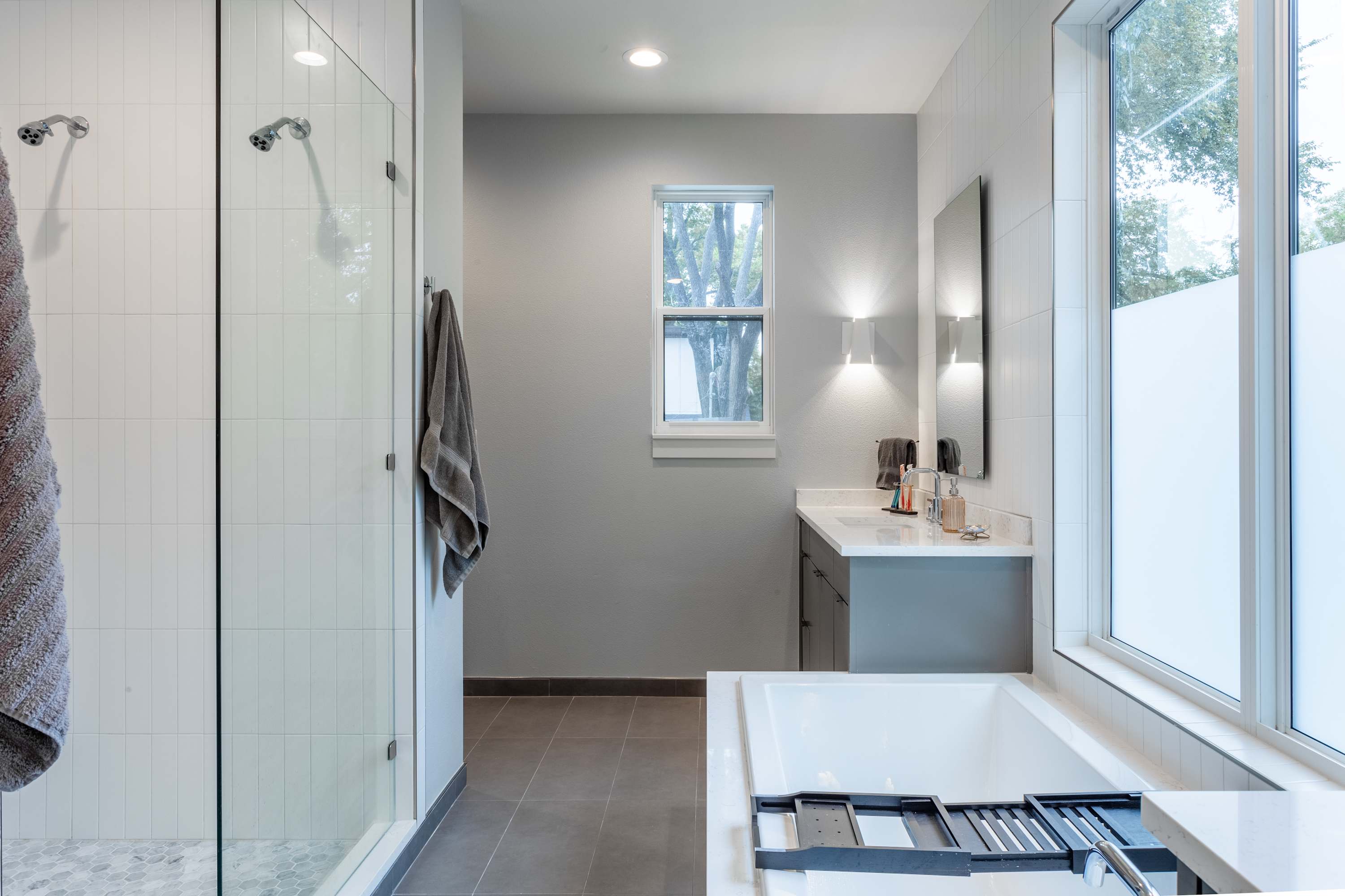 Modern bathroom with a glass-enclosed walk-in shower featuring white subway tiles and a pebble floor, alongside a built-in white bathtub and a gray vanity with a white countertop. Large windows provide natural light.