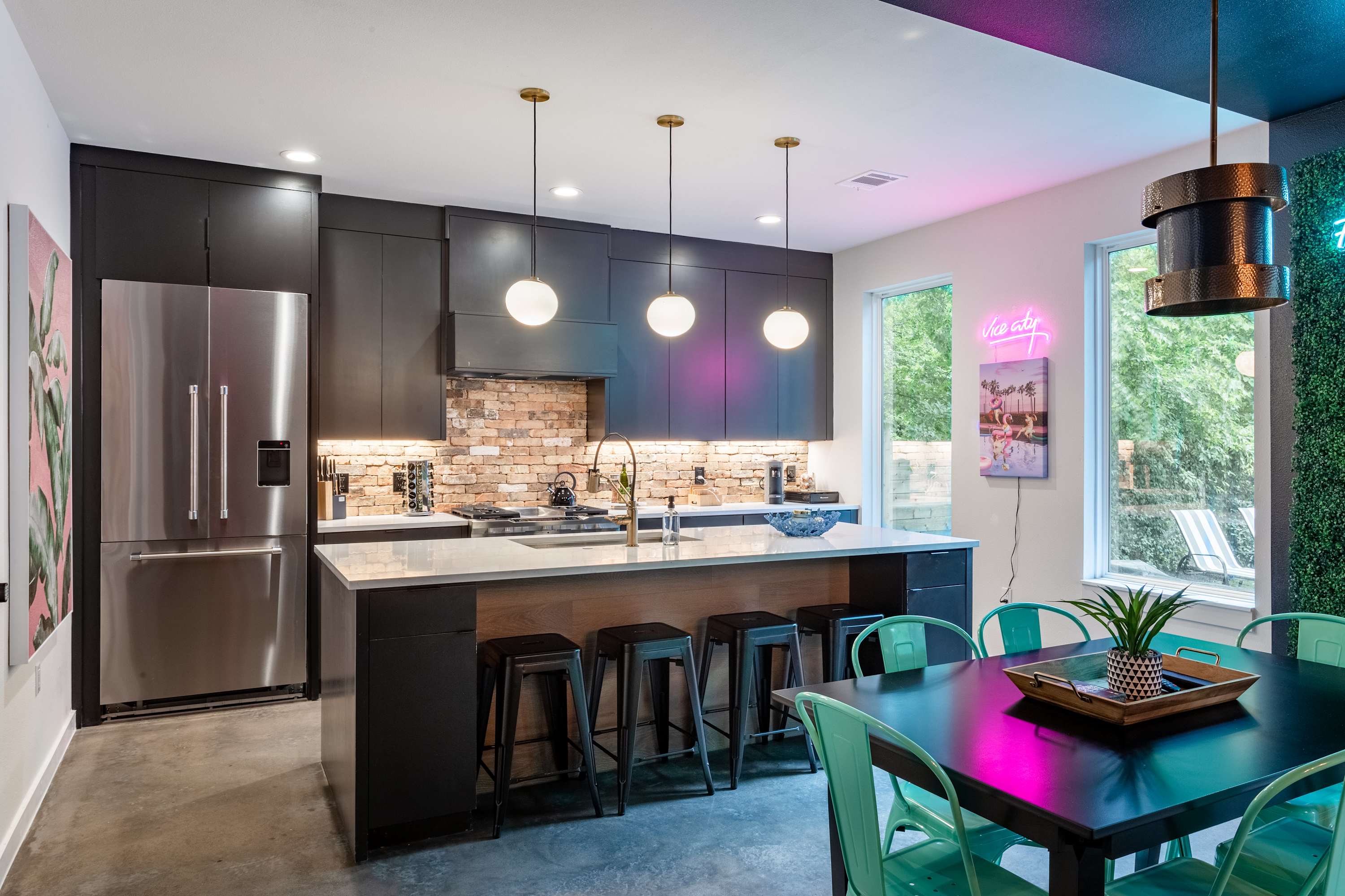 Modern kitchen with dark cabinetry, a brick backsplash, and a large island with white quartz countertops and black bar stools, flowing into a dining area with a black table and bright teal chairs. Neon signs add a vibrant touch.