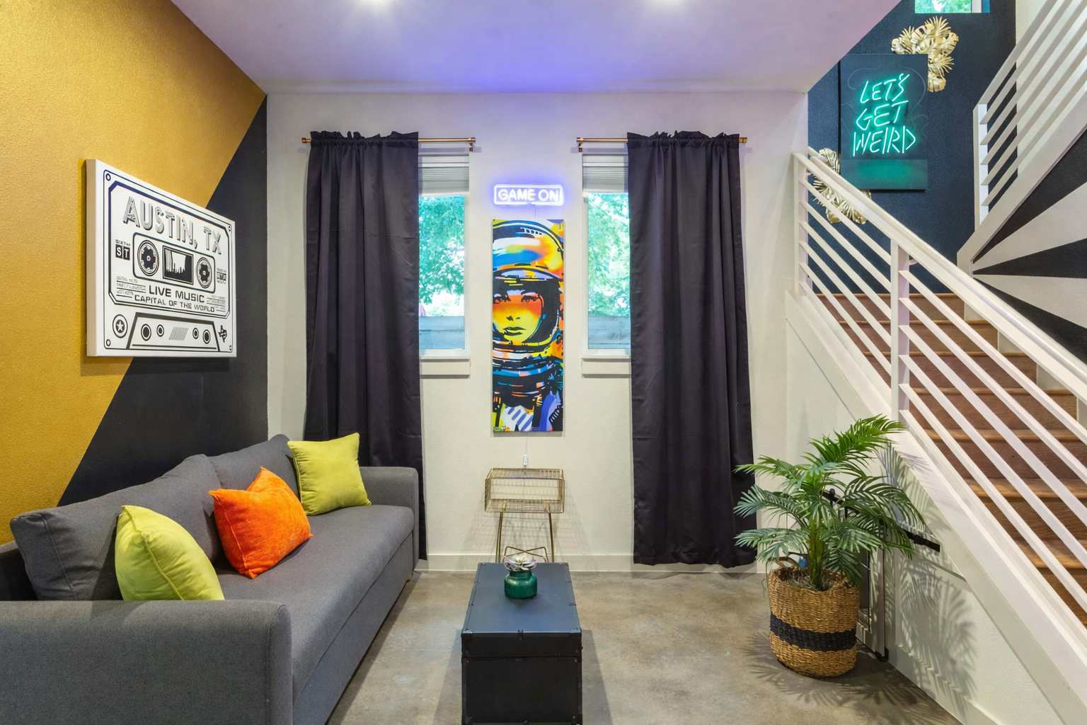 A cozy lounge area features a grey sofa with colorful pillows, a black coffee table, and a striking "GAME ON" neon sign above a vibrant astronaut artwork. The wall has a bold black and gold geometric paint design.