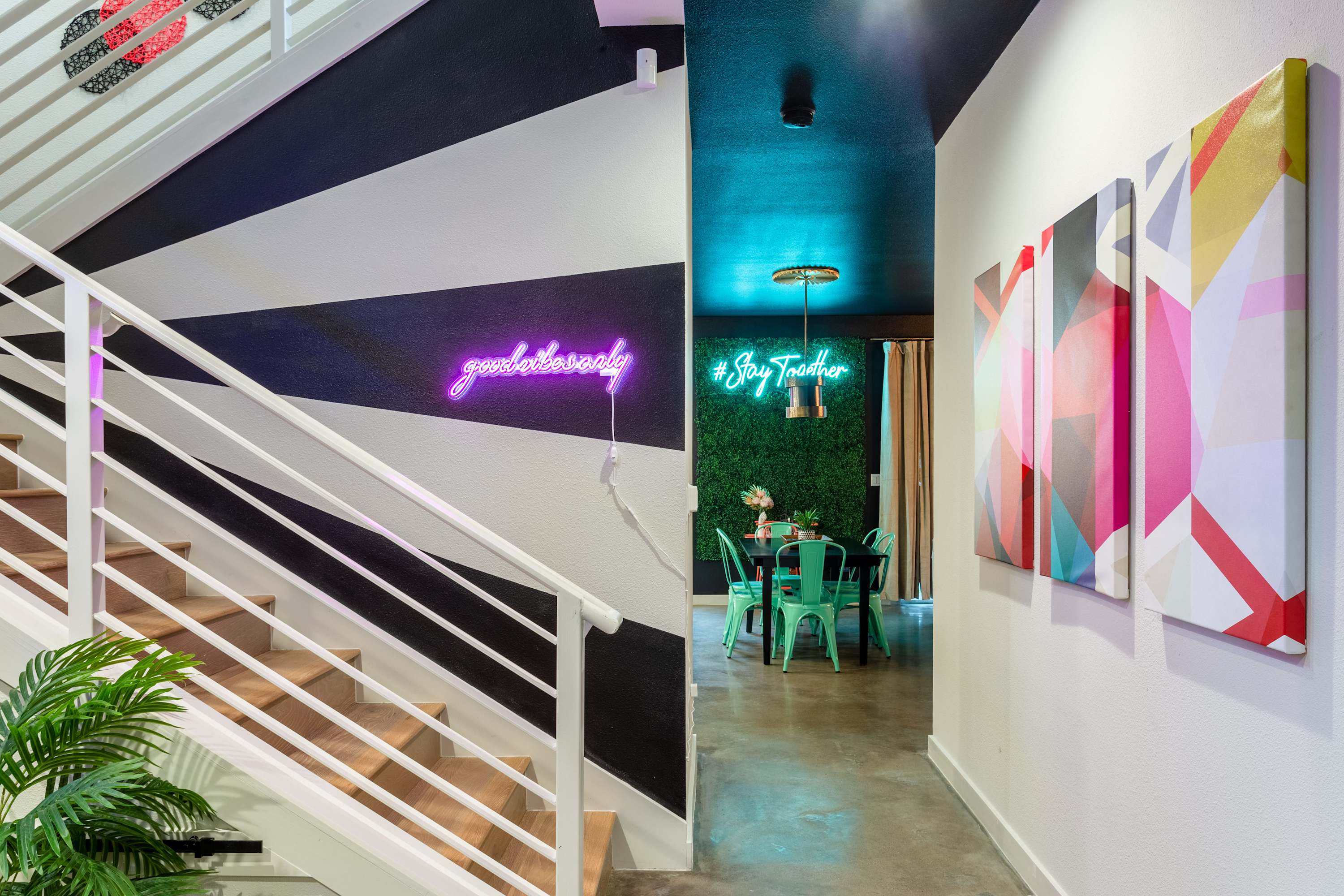 A modern staircase with white risers and wooden treads features a bold black and white geometric wall design, leading to a dining area with a green plant wall and neon signs. Colorful abstract art adorns the hallway.