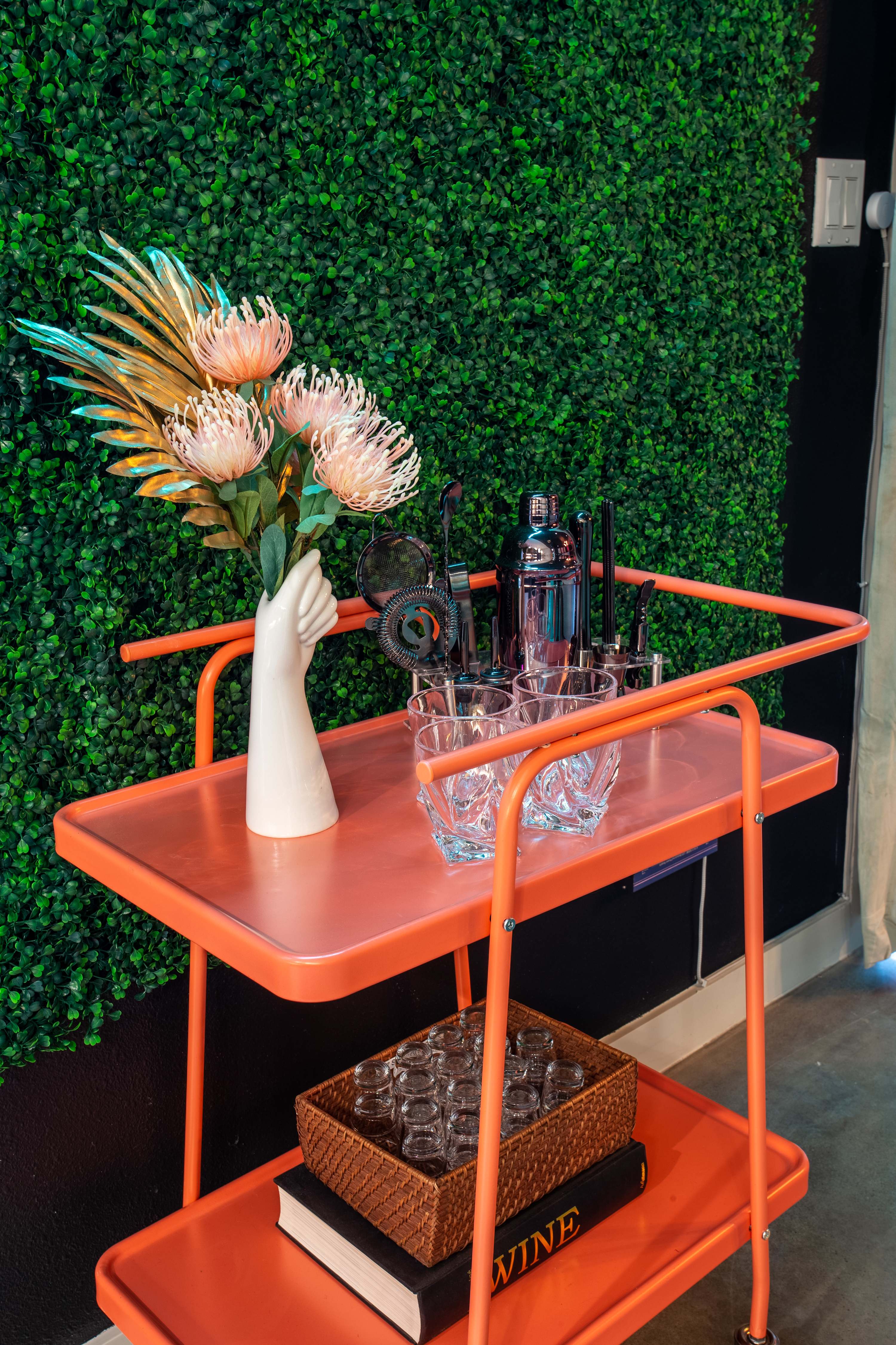 A vibrant orange bar cart stands against a lush green faux plant wall, adorned with a white vase holding exotic flowers and various cocktail-making tools. Shot glasses are neatly arranged in a basket on the lower shelf.