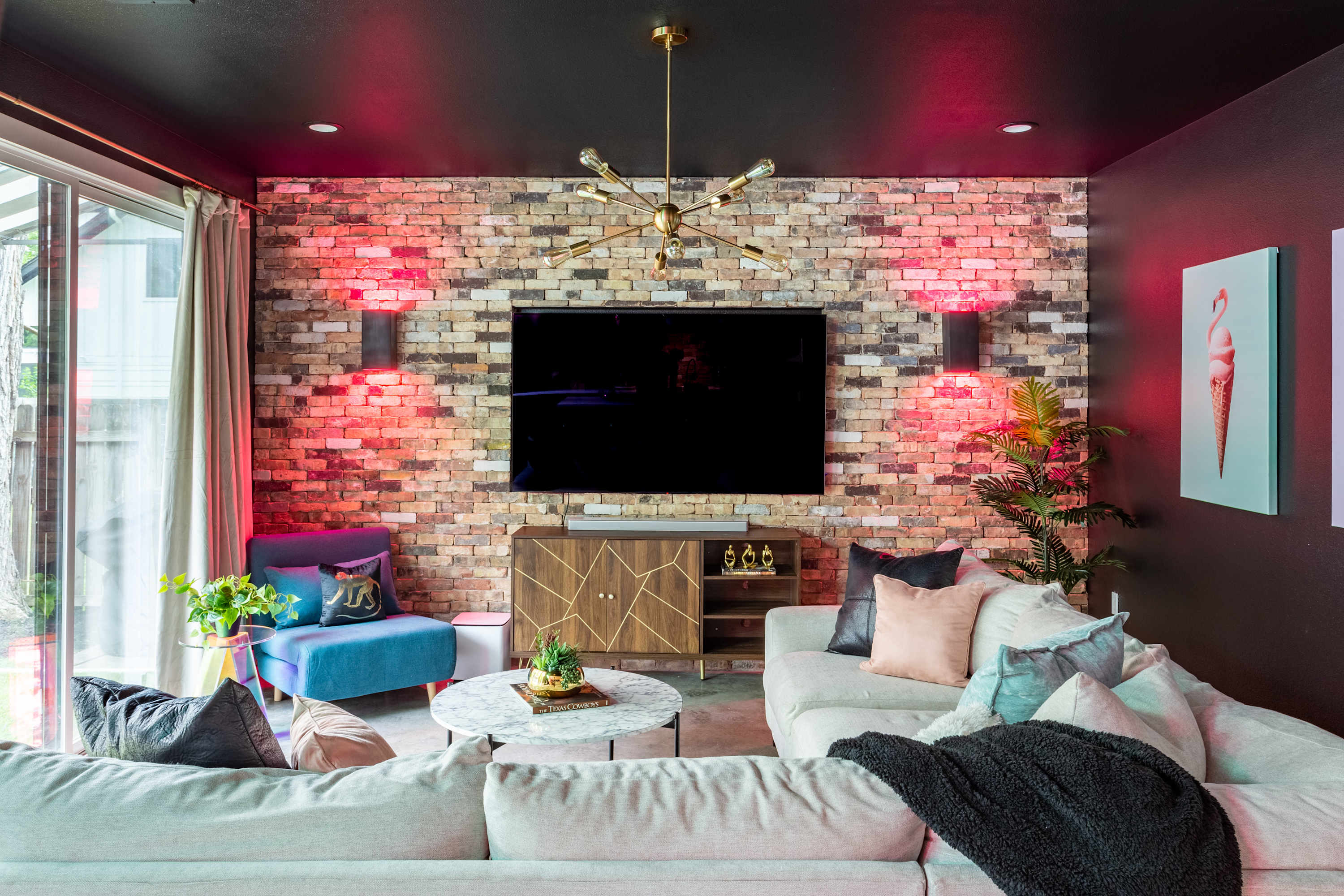 Vibrant living room with a large sectional sofa, a brick accent wall illuminated by red uplighting, and a large wall-mounted TV above a wooden console. A gold sputnik chandelier and eclectic decor complete the space.