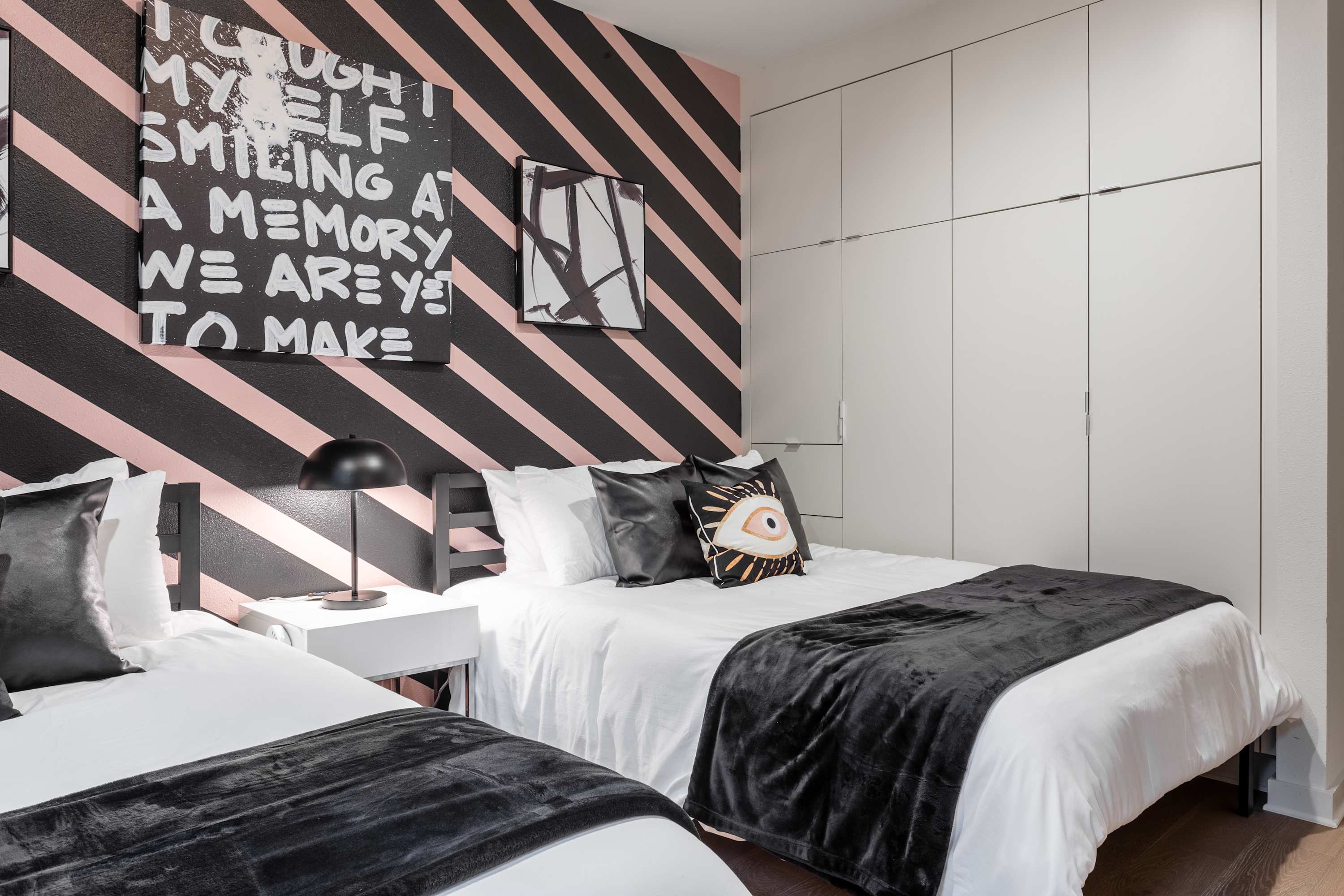 Stylish bedroom featuring two twin beds with black and white bedding, a black and pink striped accent wall, and a built-in white closet. A large abstract artwork and a black table lamp complete the modern aesthetic.