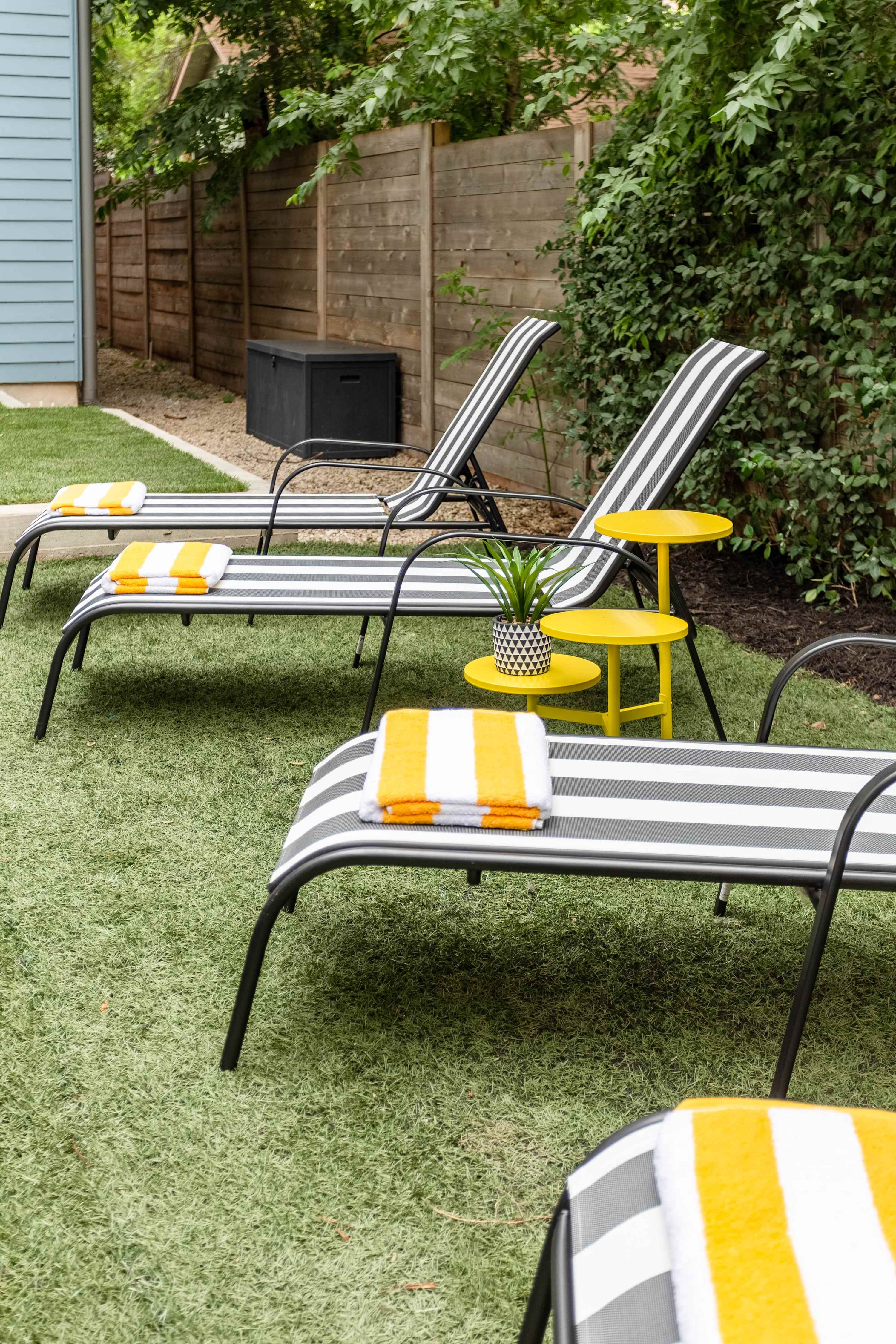 A close-up of three black and white striped lounge chairs on artificial turf, each with a yellow and white striped towel. Yellow side tables are placed between the chairs, and a wooden fence is in the background.