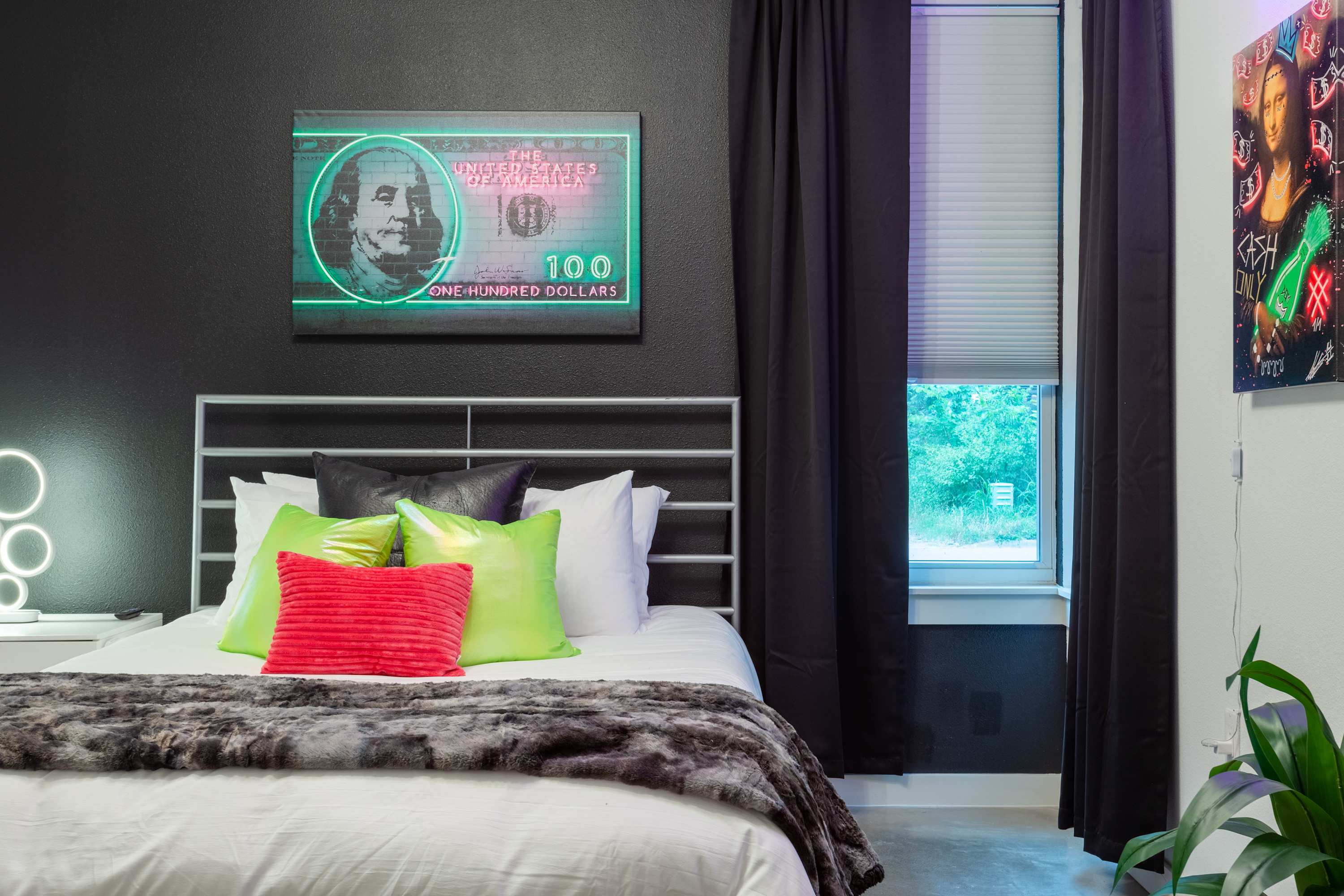 A close-up of a modern bedroom showcasing a white metal bed with vibrant green and pink pillows, a faux fur throw, and a striking neon-lit Benjamin Franklin artwork on a dark accent wall. A white nightstand with a unique lamp is visible.