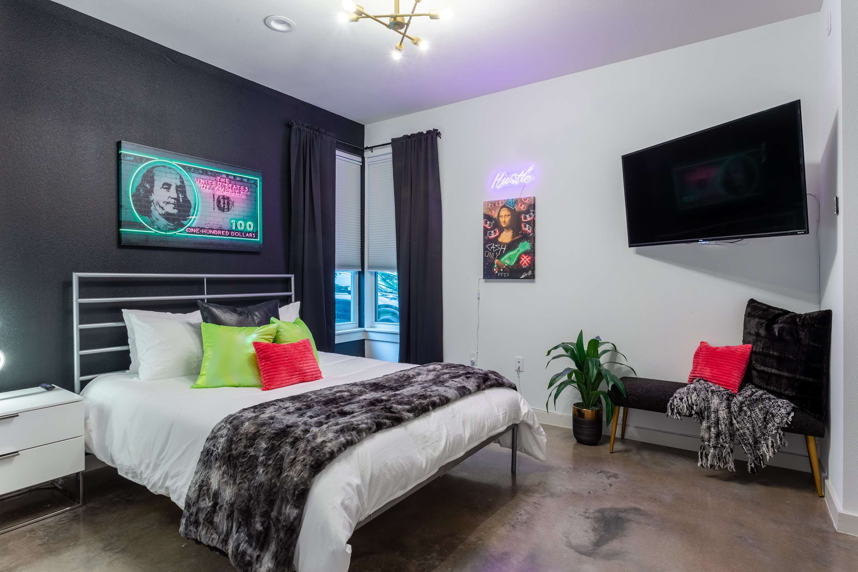 A stylish bedroom features a white metal bed frame with a faux fur throw and bright green and pink pillows, set against a dark accent wall with a neon-lit Benjamin Franklin artwork. A wall-mounted TV and a bench complete the space.
