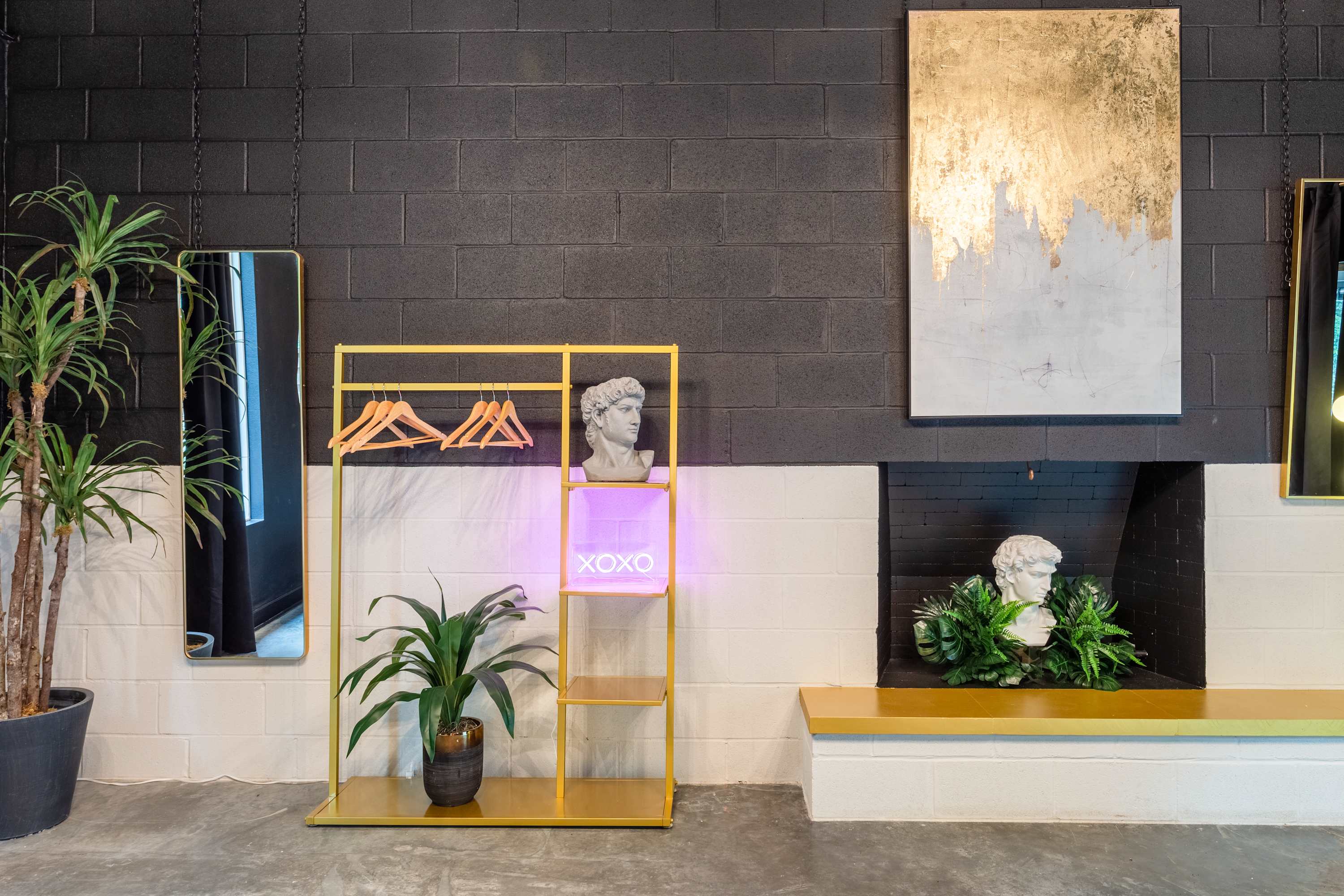 A stylish corner of a common area featuring a gold clothing rack with shelves, a vibrant neon 'XOXO' sign, and classical bust sculptures. The walls are black painted cinder block with a white wainscoting, accented by a large abstract gold and white painting.