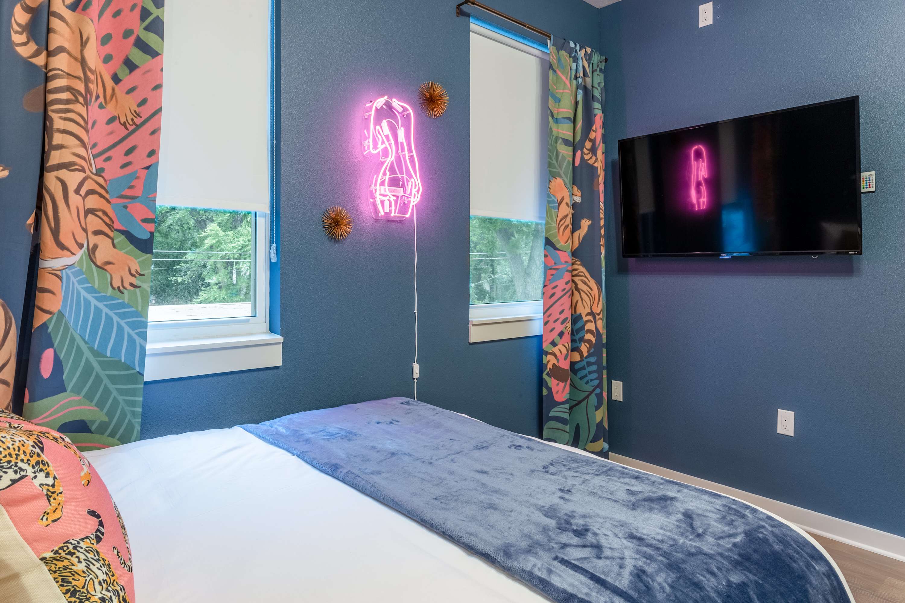 Colorful bedroom featuring a queen bed with blue throw and leopard print pillows, a wall-mounted TV, and tropical print curtains. A pink neon high-heel sign illuminates the blue wall, complementing the vibrant decor.